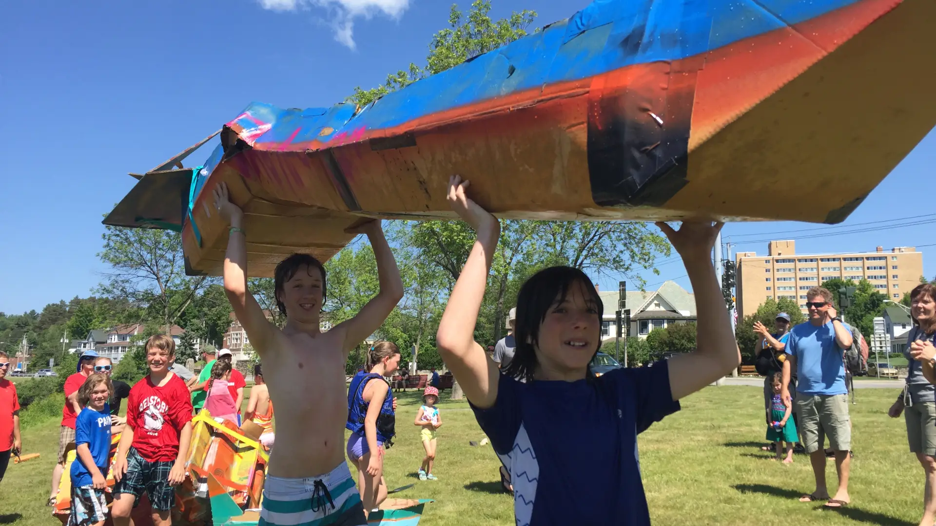 two boys holding a cardboard boat over their head as other look on in the background 
