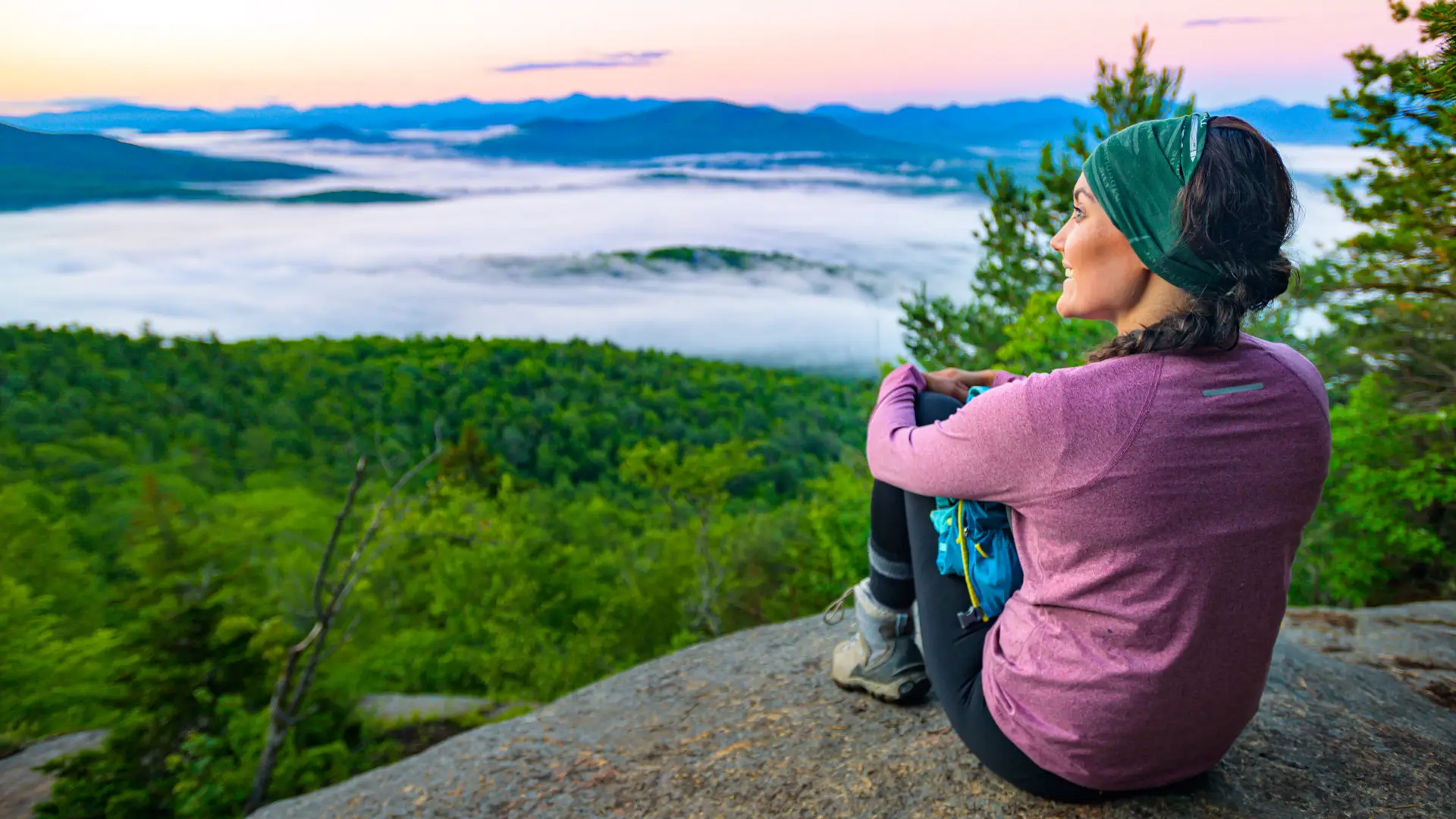 Woman in a purple shirt sits on the edge of Baker Mountain and stares out overtop of the green trees below with light cloud cover and other Adirondack mountains in the distance