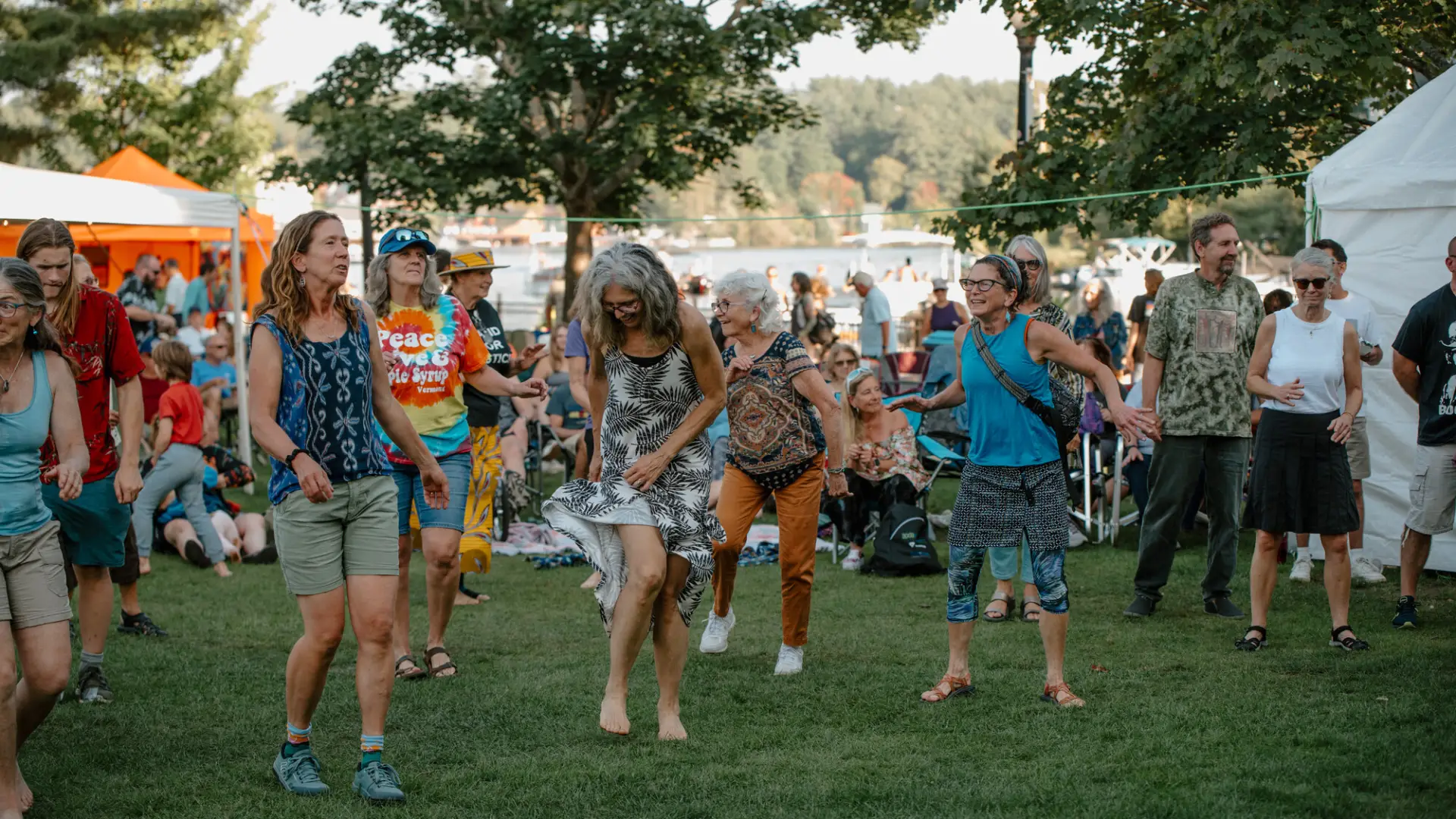 A group of people dancing in the grass surrounded by families and vendor tents on the green by Lake Flower in Saranac Lake.