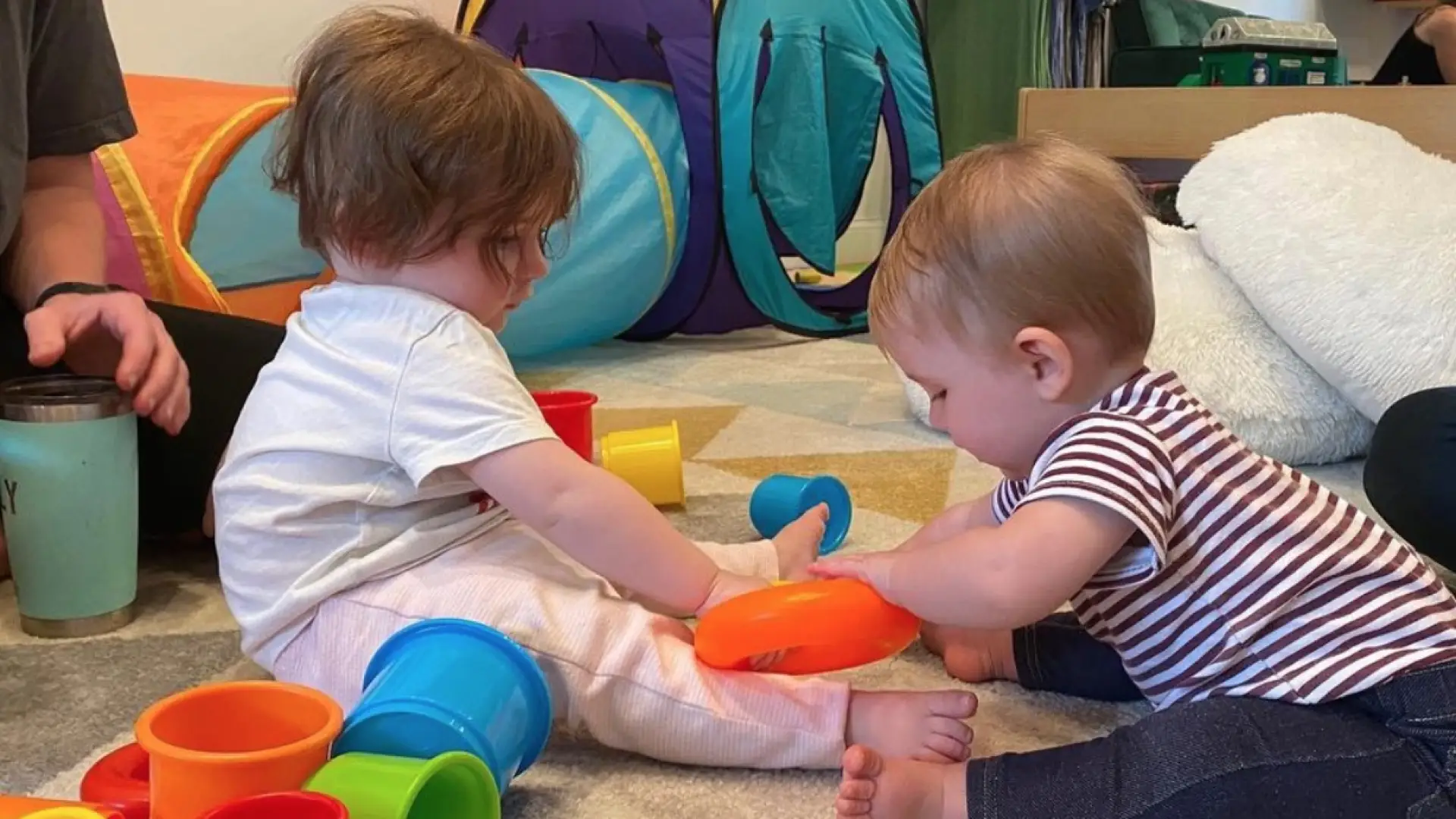 Babies sit and play with colorful stacking cups on a floor together