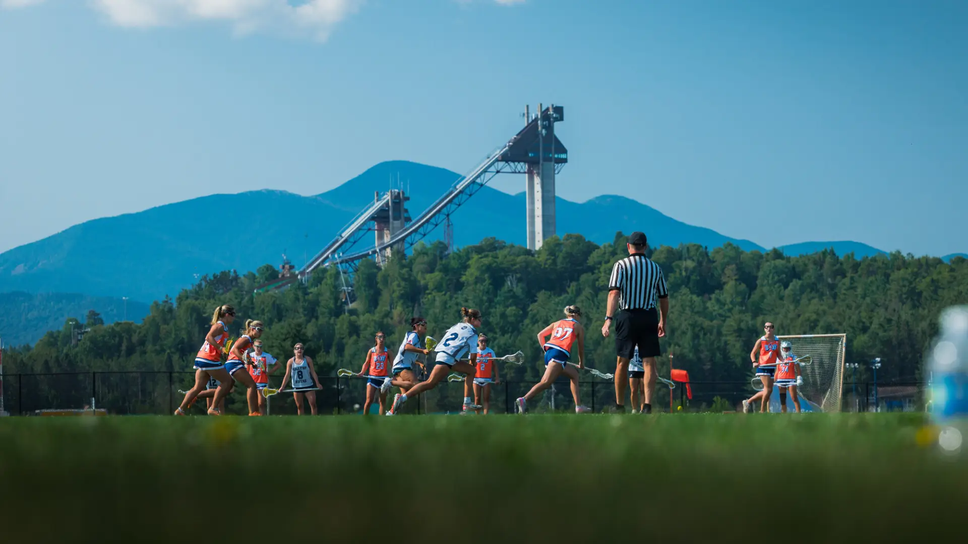 Two teams of women play lacrosse on field in front of mountains and ski jumps while a referee looks on