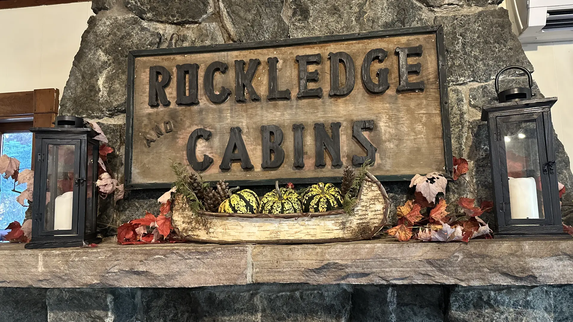 A carved sign reading 'Rockledge and Cabins' over a stone mantle with birch bark boat filled with festive squash and candles.