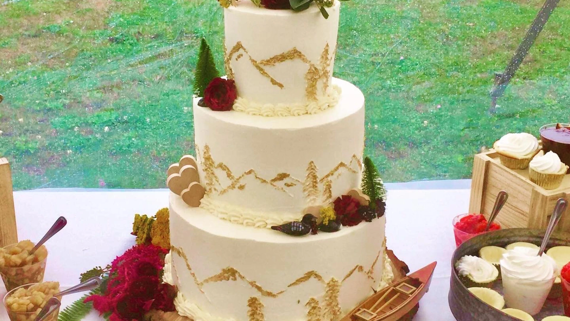 A three tiered cake frosted with an abstract mountain design and topped with adirondack chairs.