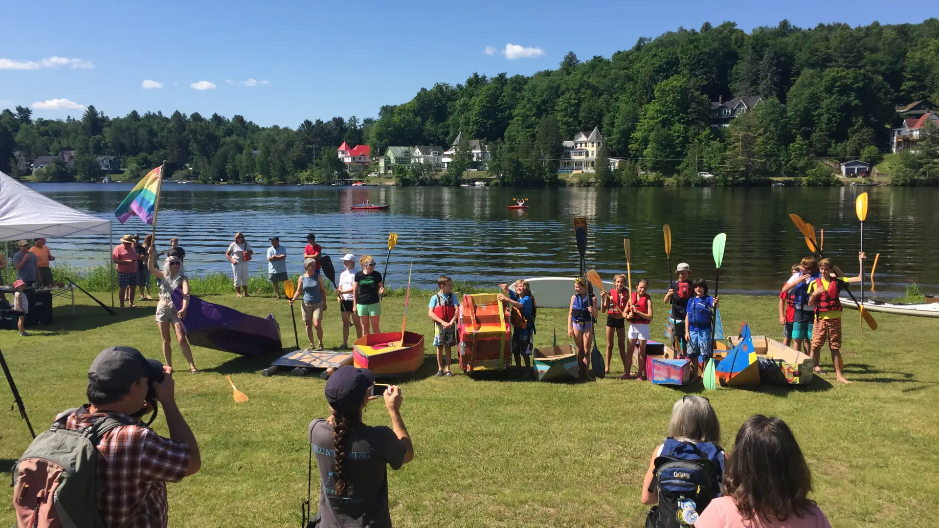 group of people at the lake shore with cardboard boat materials and oars, getting ready to go in the lake 