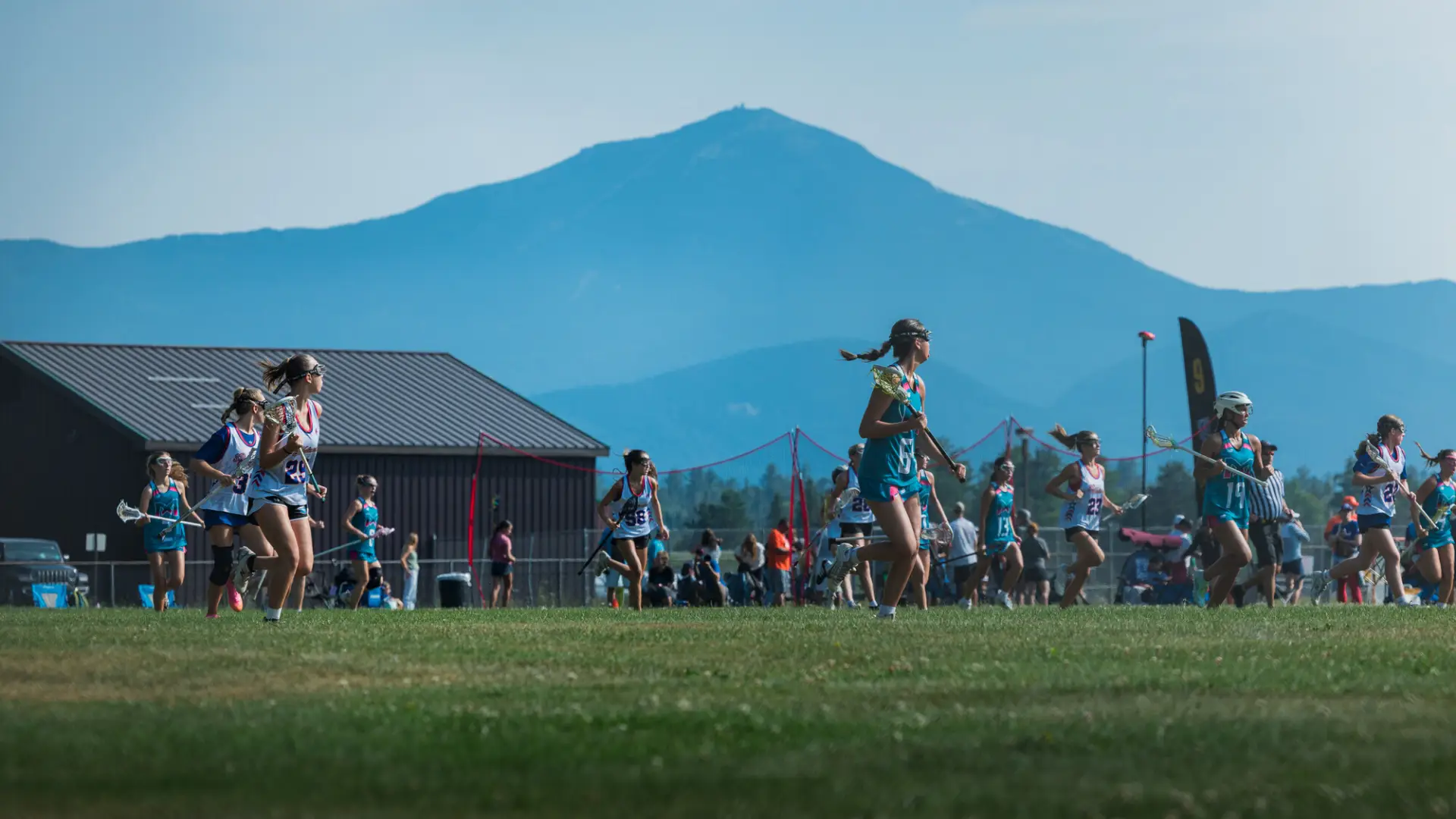 Female lacrosse players from two teams run down field with Whiteface Mountain in background