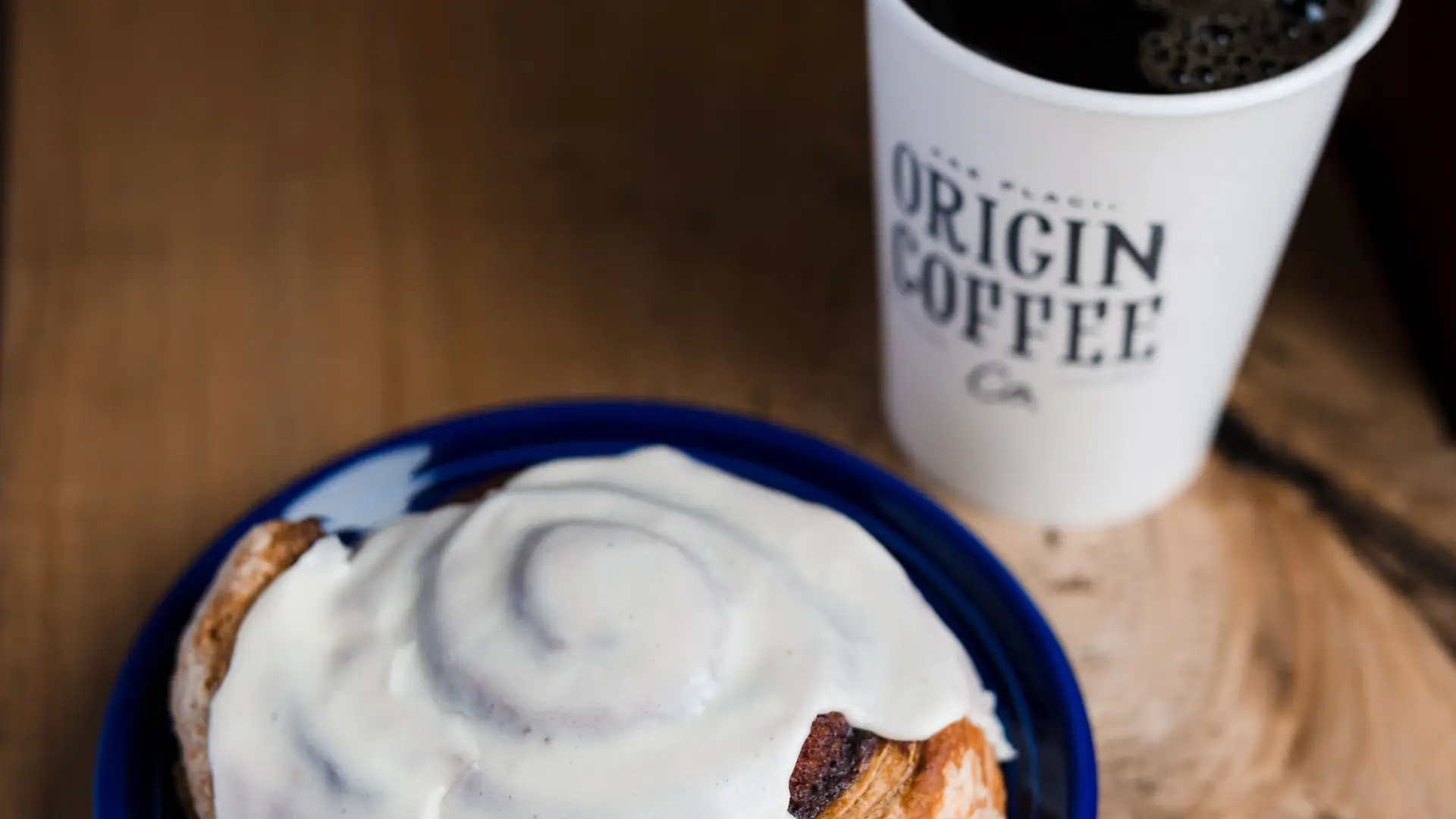 A large cinnamon bun, smothered in icing on a blue plate next to a black drip coffee in a branded Origin coffee cup.