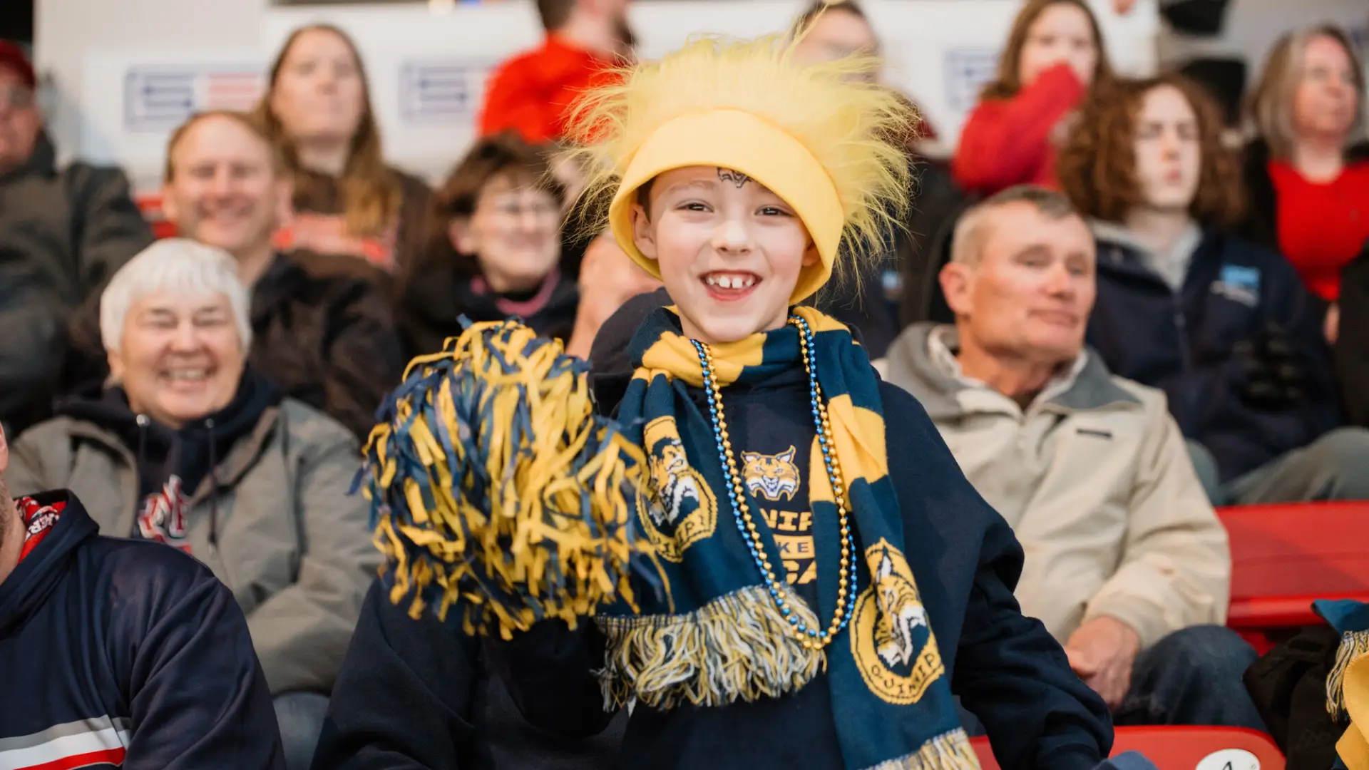 Child dressed in Quinnipiac hockey gear and accessories looks at camera and smiles while cheering in the stands.