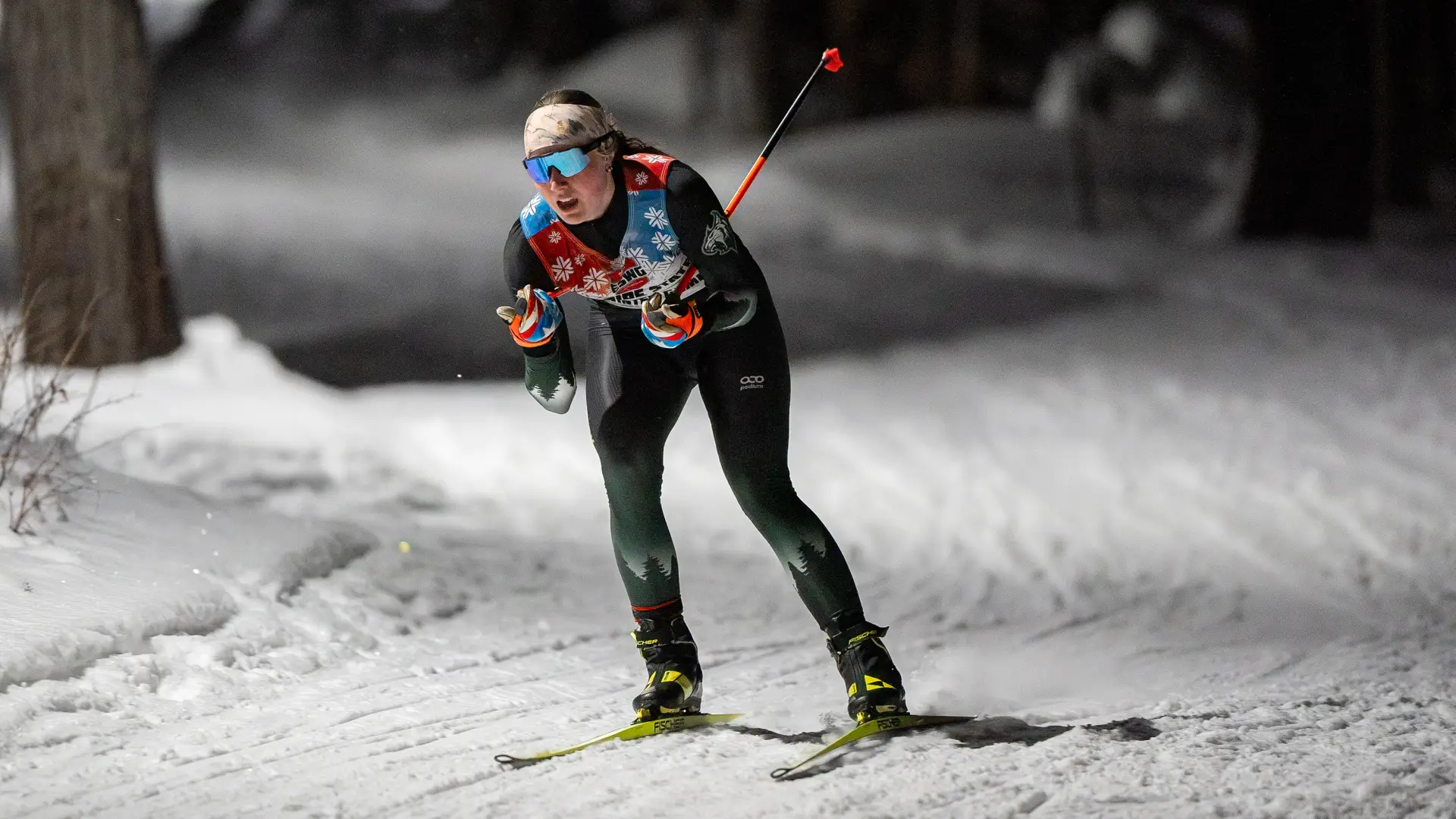 A cross-country skier in a racing suit and sunglasses glides along a snowy trail at night under bright artificial lighting.