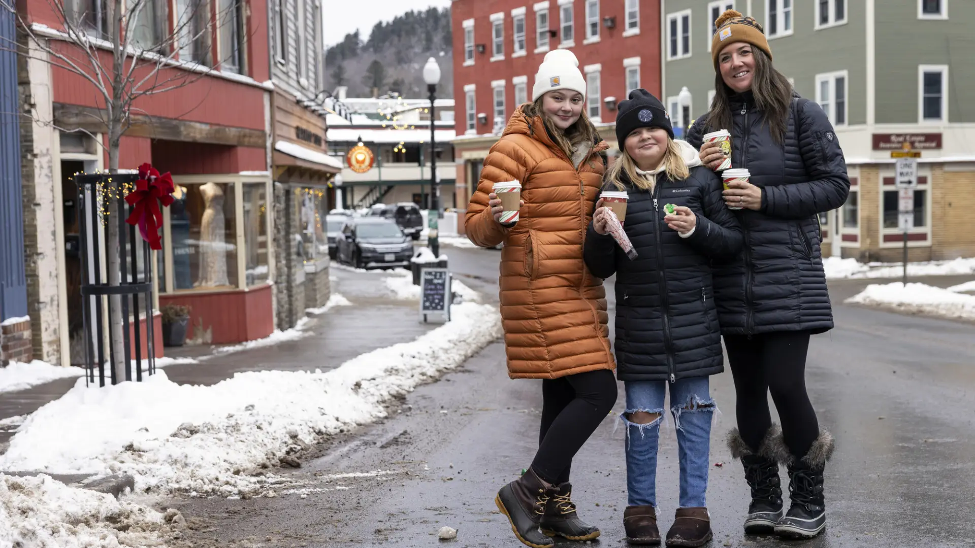 Three women standing in the middle of a winter village smile with hot cocoa in their hands. 