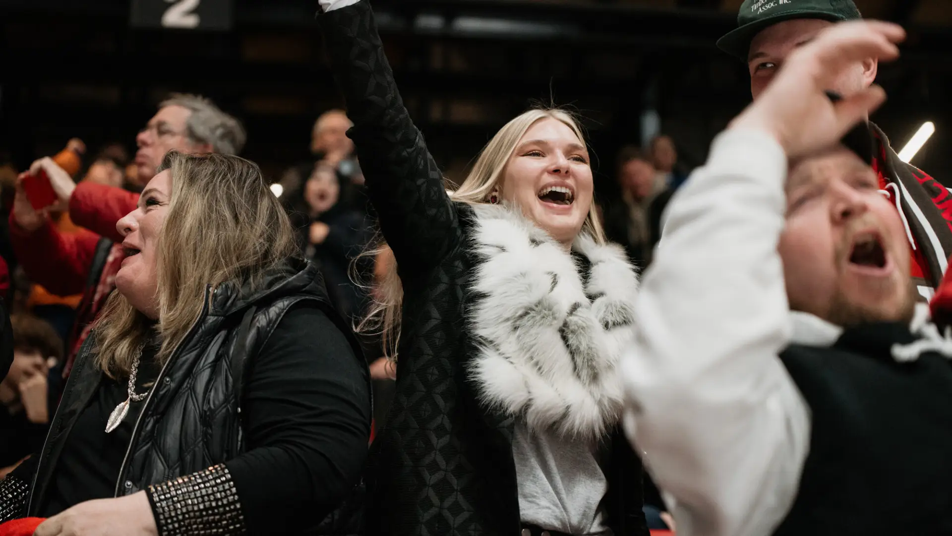 Woman dressed in black with furry white scarf holds arm above her head while cheering in the stands.