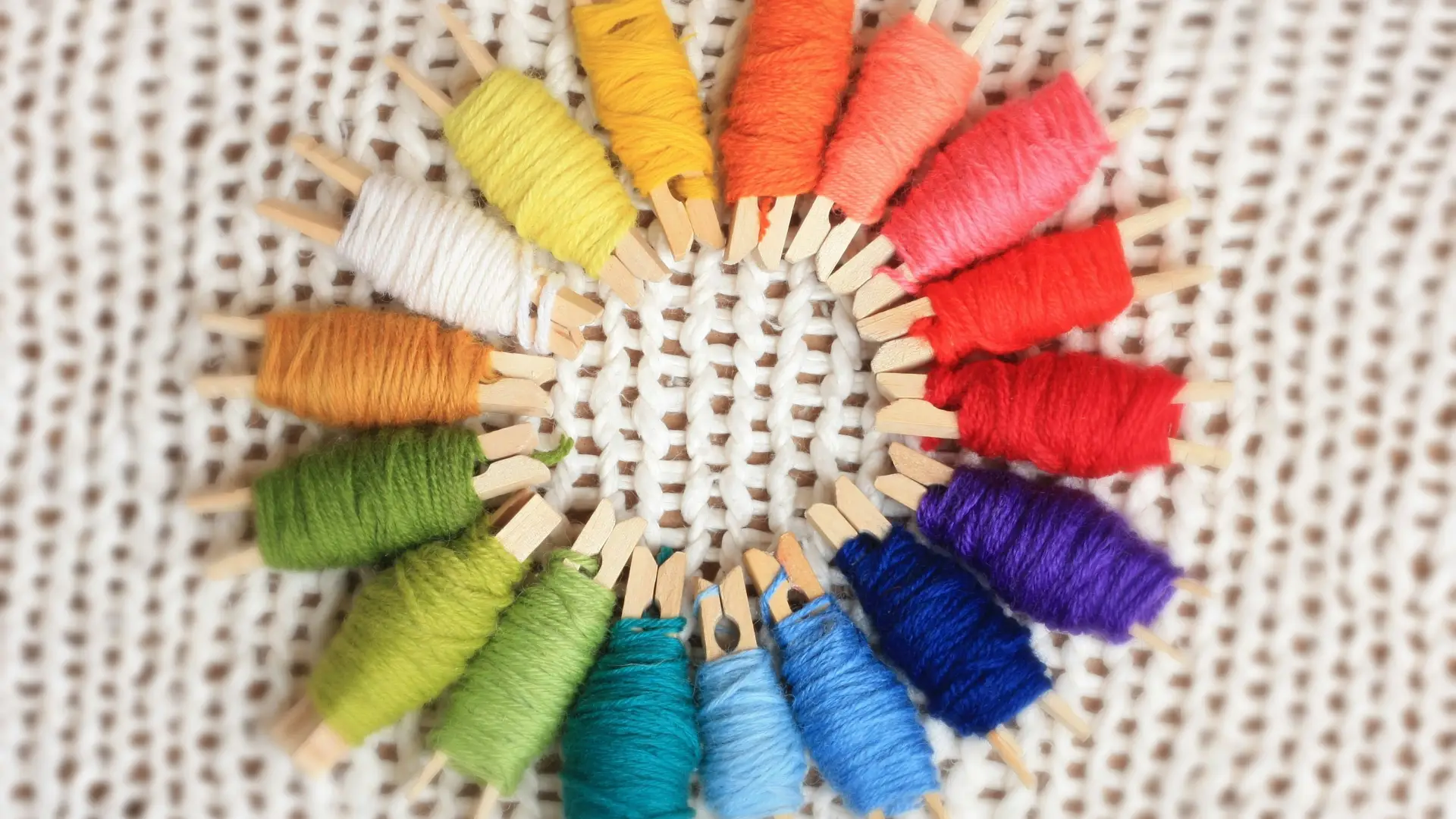 A view of colorful yarns wrapped around wooden clothespins, arranged in a rainbow wheel on a white knitted background.