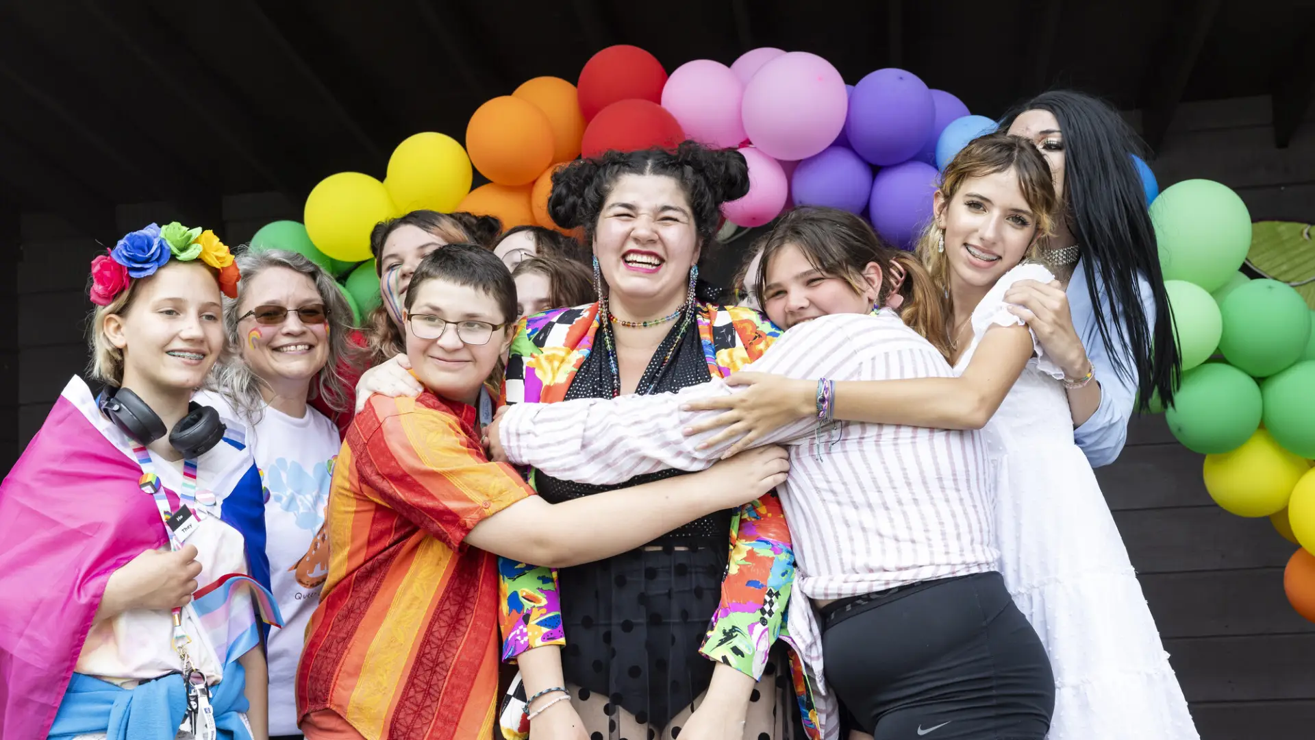 A variety of people hugging in front of a rainbow arch of balloons.
