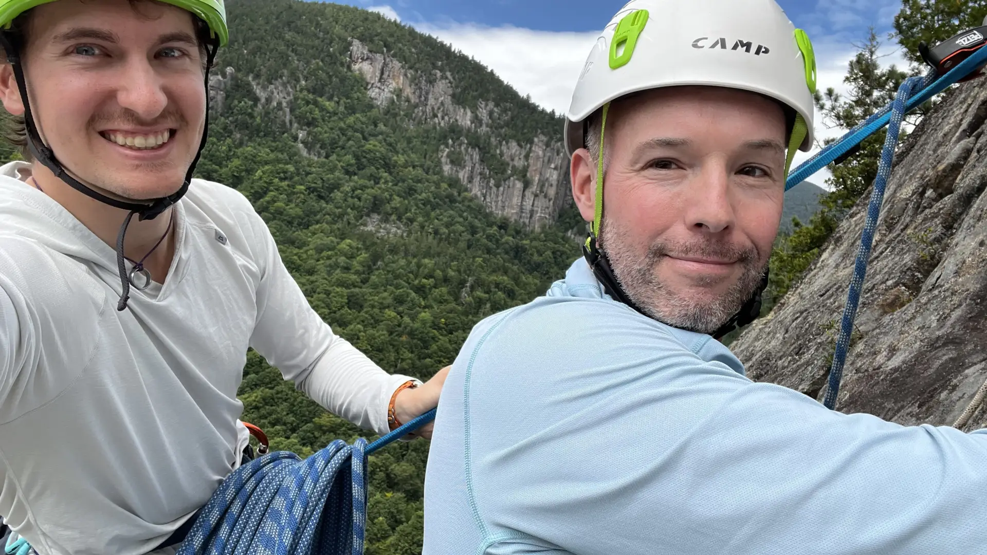 Two men smile into the camera while climbing a rock ledge