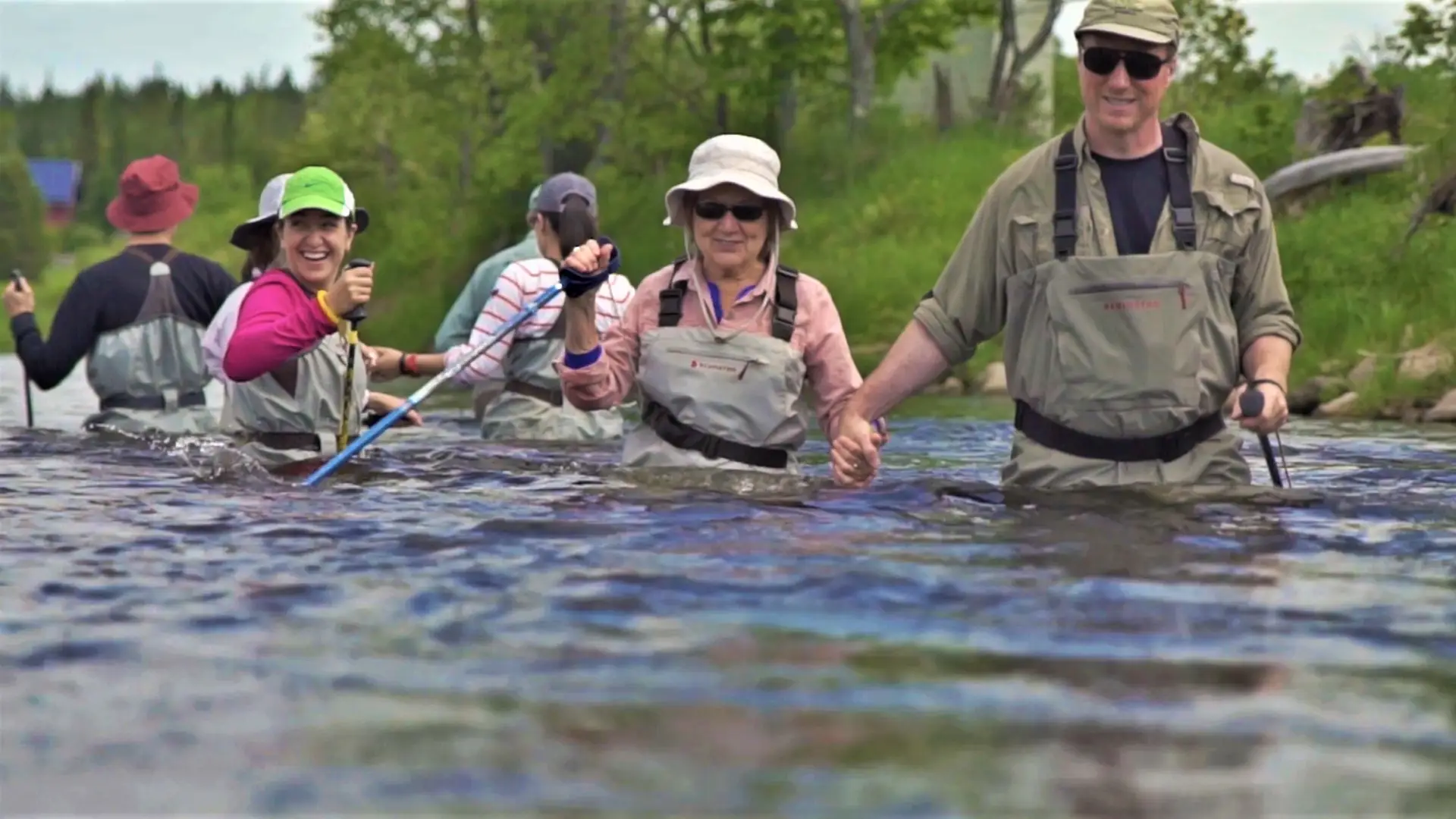 A group of people in chest wadders and walking sticks journey through a river