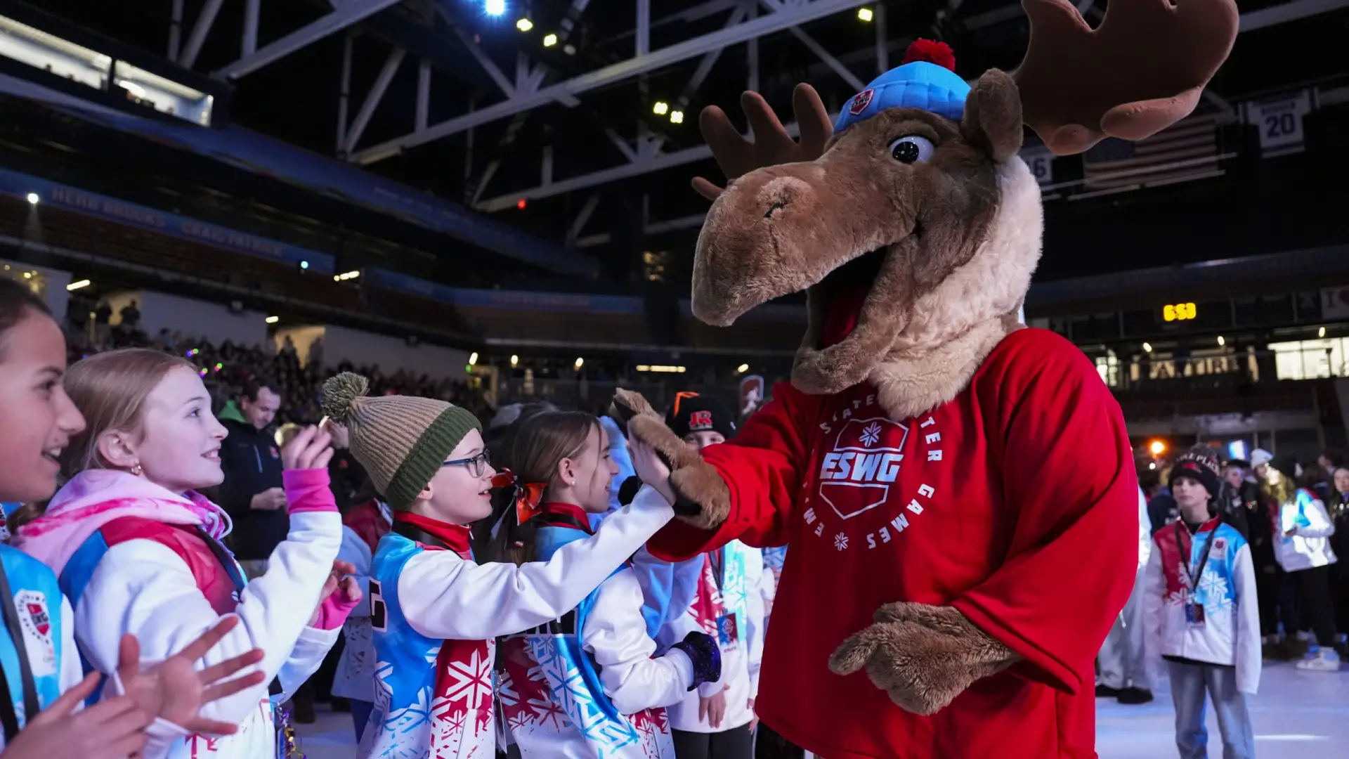 A group of children in colorful winter jackets high-five a person in a cheerful moose mascot costume.
