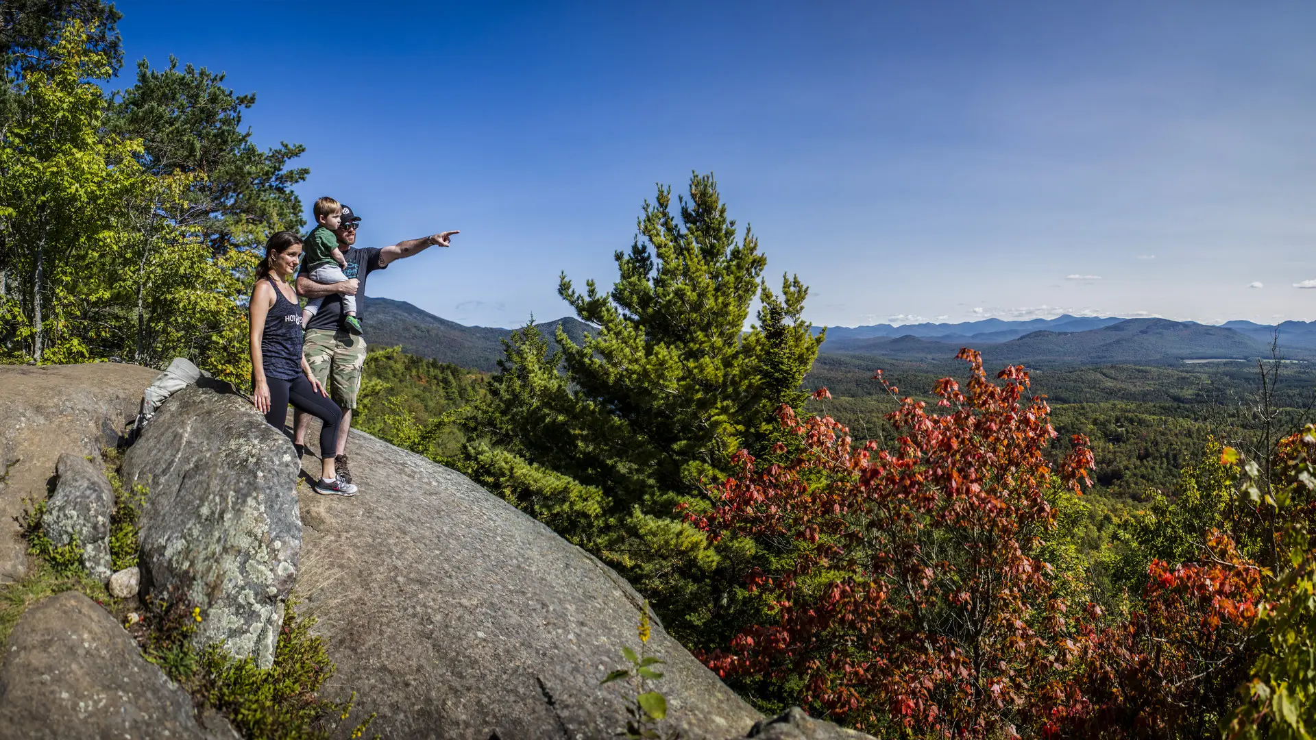 A couple holding a toddler points off into the distance from the top of Baker Mountain with some of the trees closest to them starting to turn red