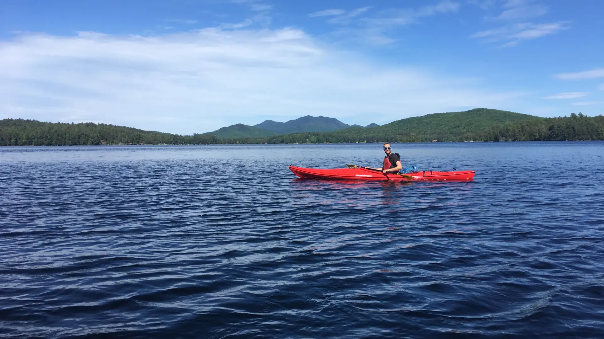 Kayaker in a red kayak paddling across Lower Saranac Lake, with mountains and blue skies in the background