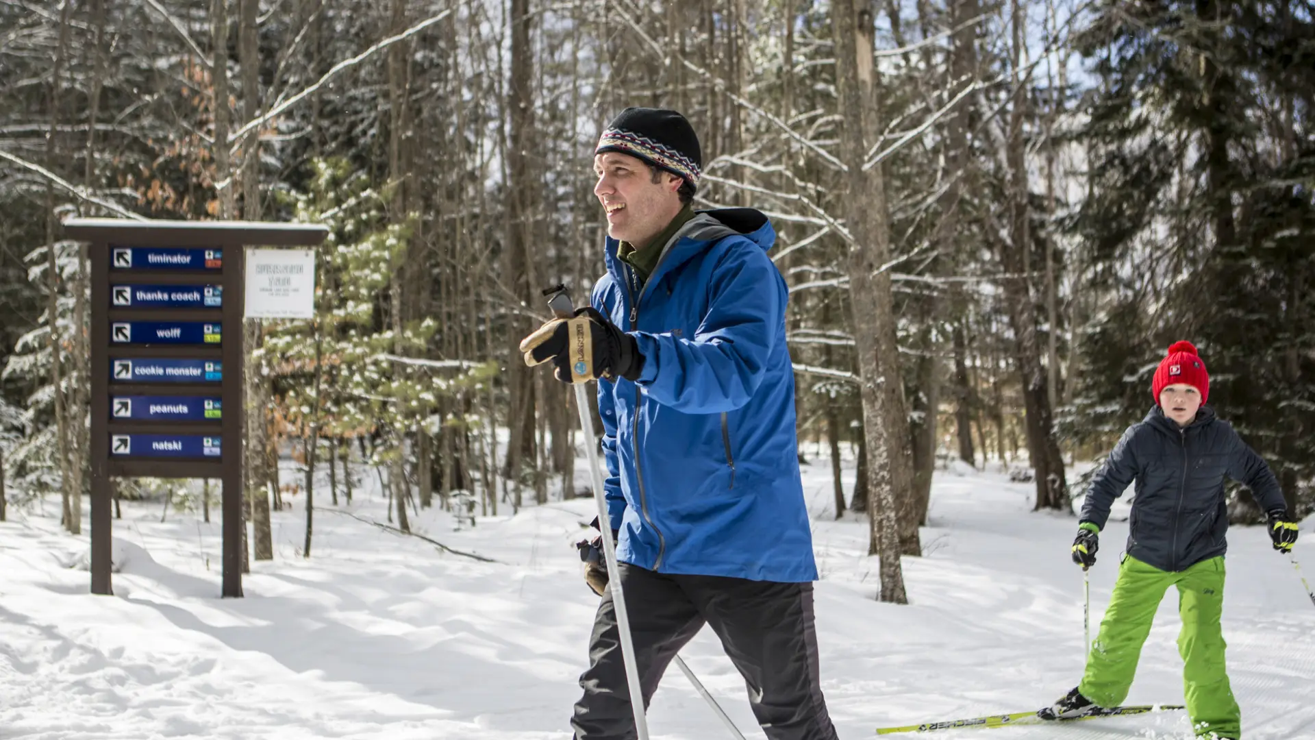 A father skis past a Dewey Mountain trail sign with a happy child following.