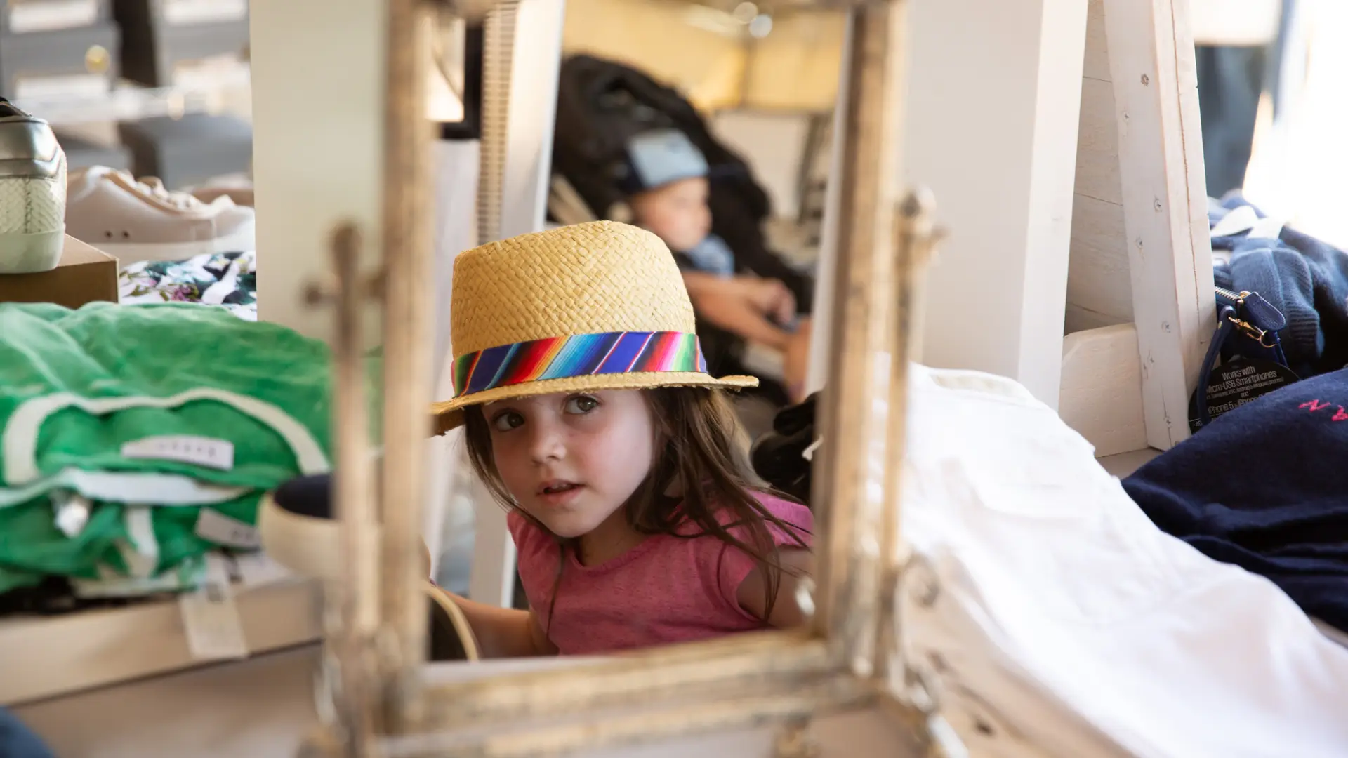 A little girl checks her self out in a mirror while wearing a straw hat with rainbow ribbon.