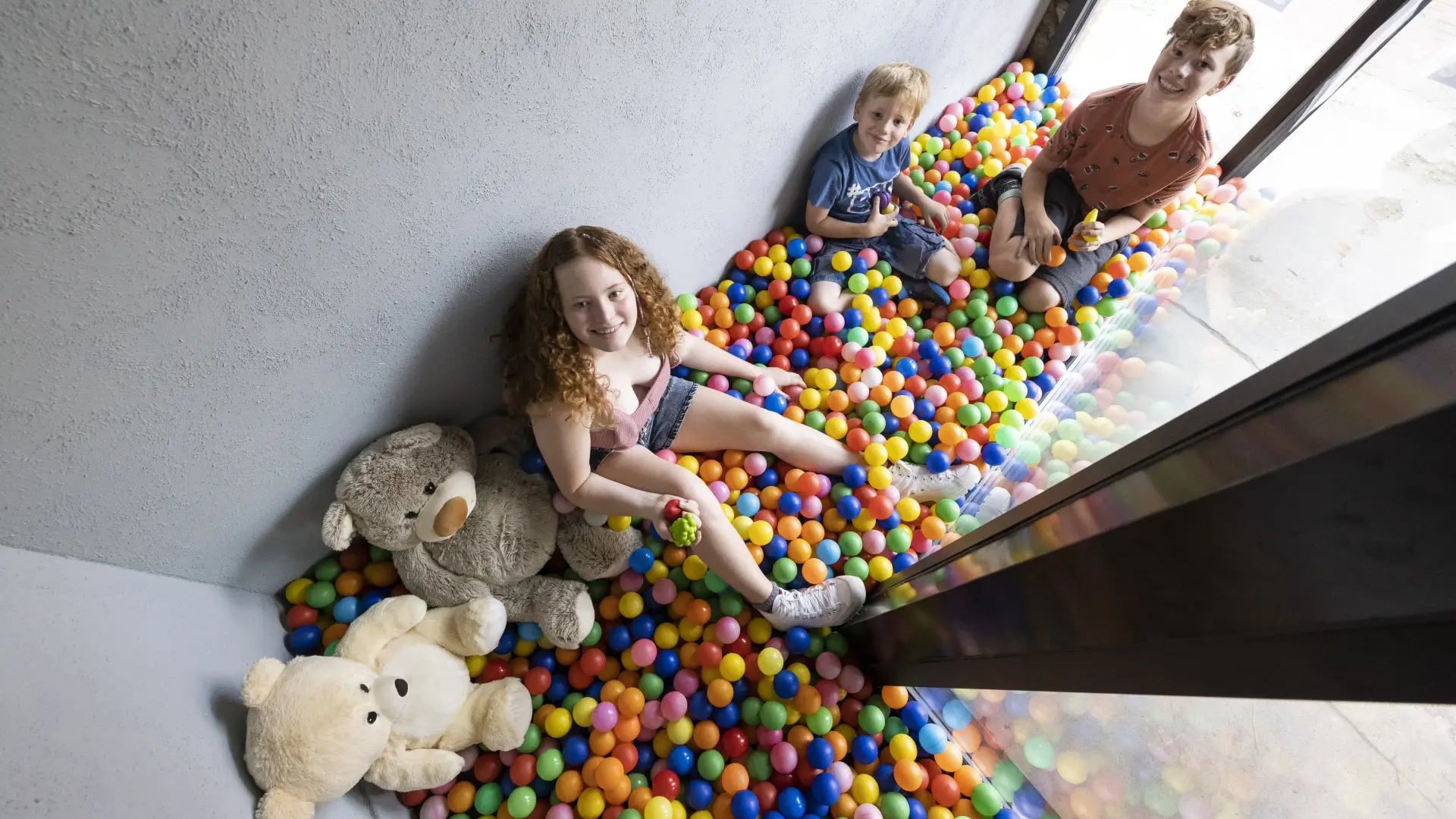 Kids smiling and playing in a colorful ball pit by windows