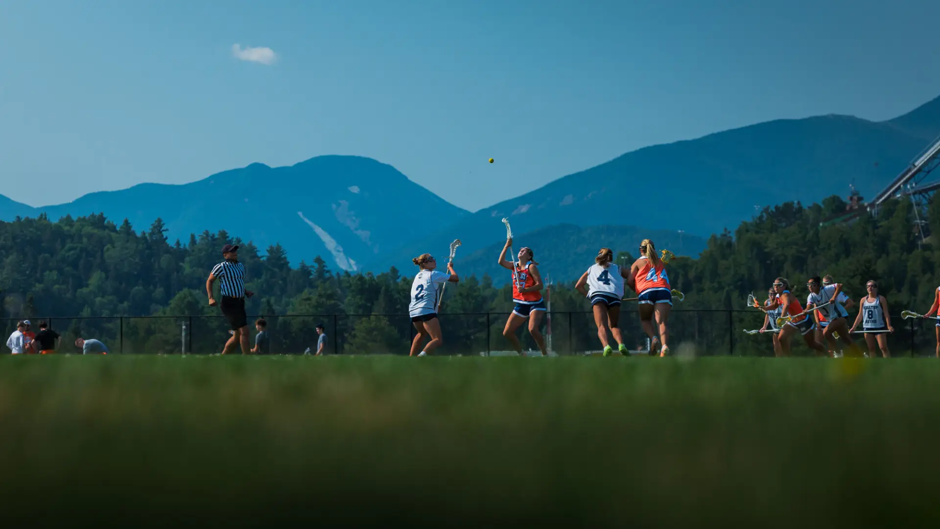 Two female lacrosse players look up at yellow lacrosse ball during play on field in front of Adirondack Mountain backdrop