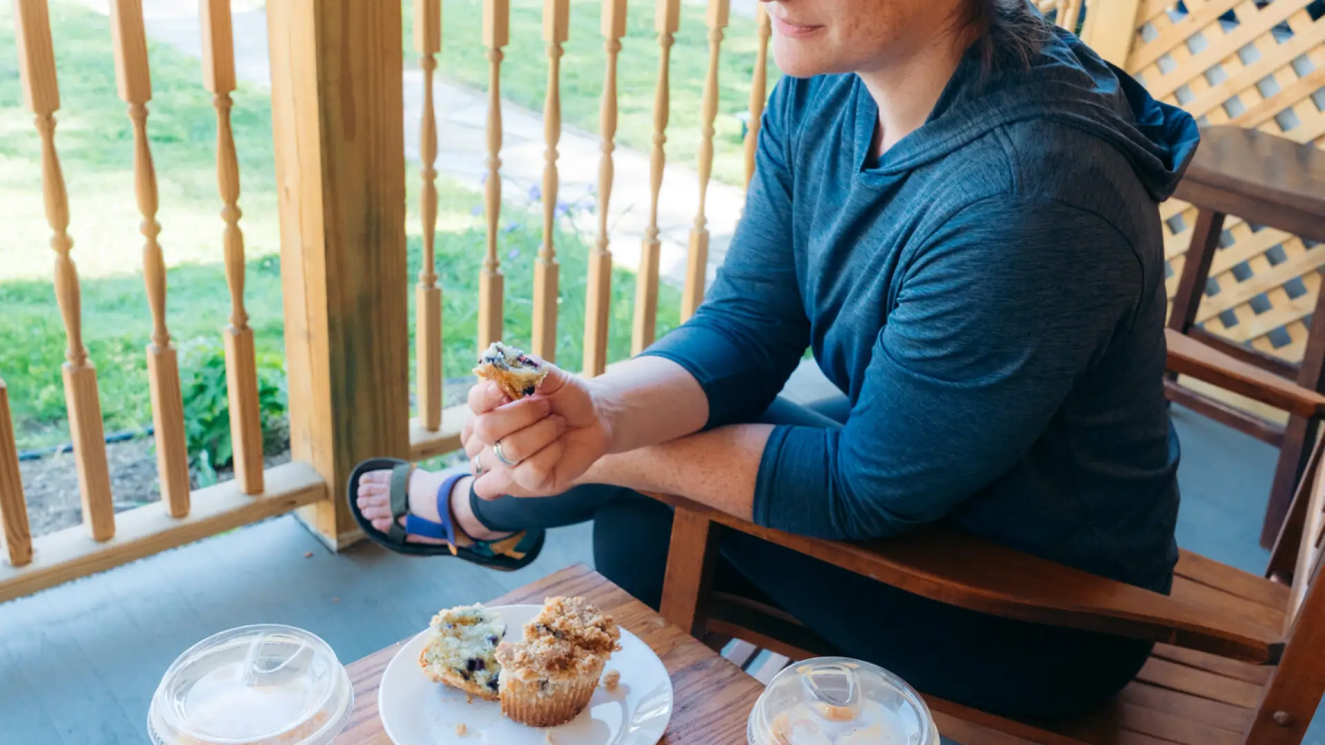 A woman sits in a rocking chair with a muffin and iced coffee on a small table next to her. 