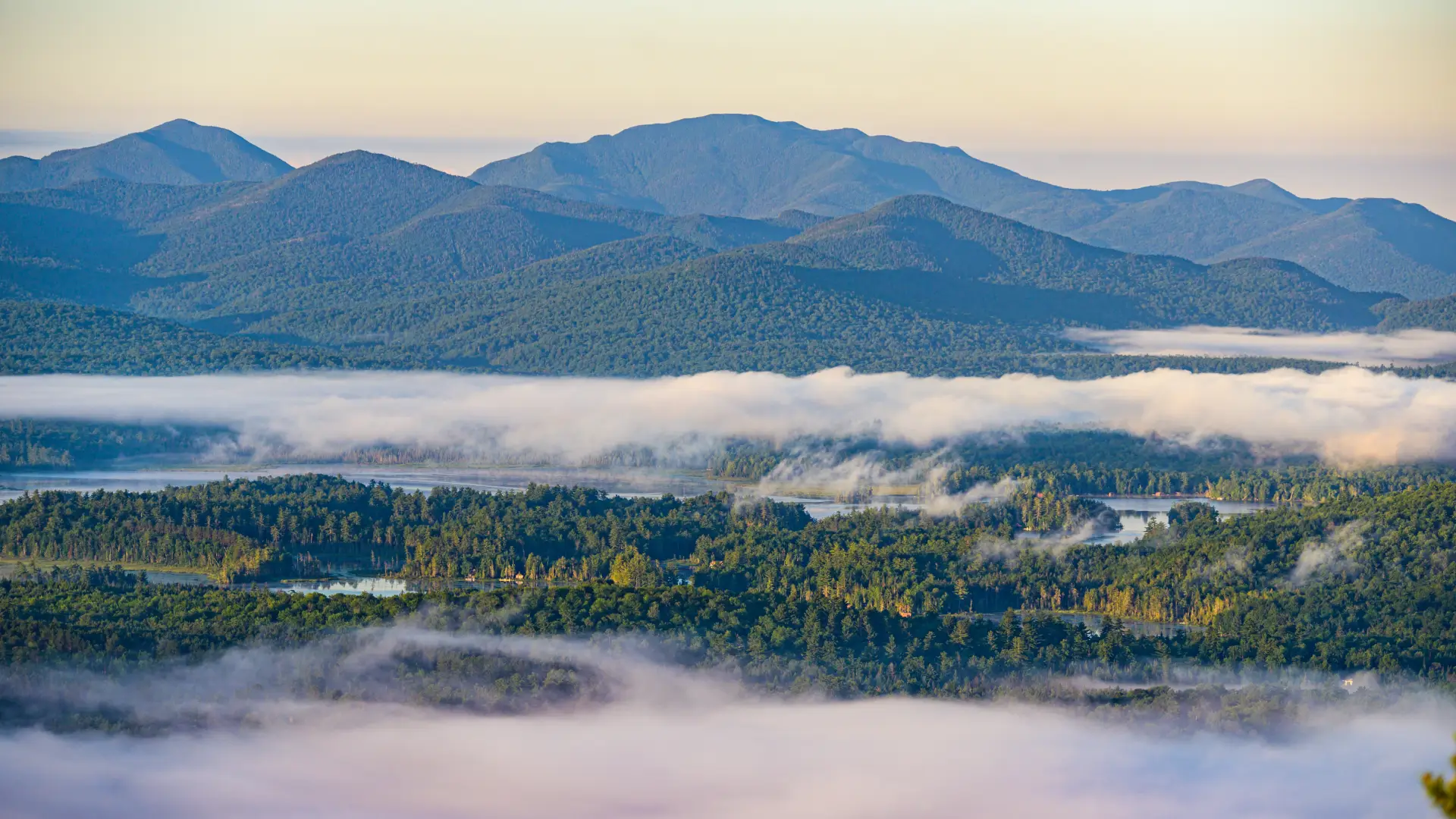 View from the top of Baker Mountain with green trees and light cloud cover below and other Adirondack mountains in the distance