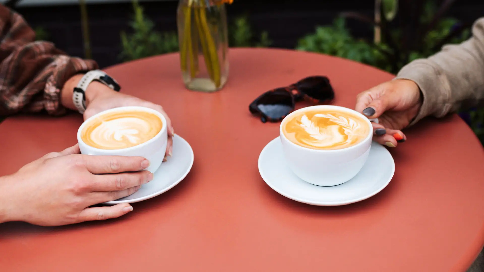 Two lattes sit on a colorful table with hands extending to retrieve them.