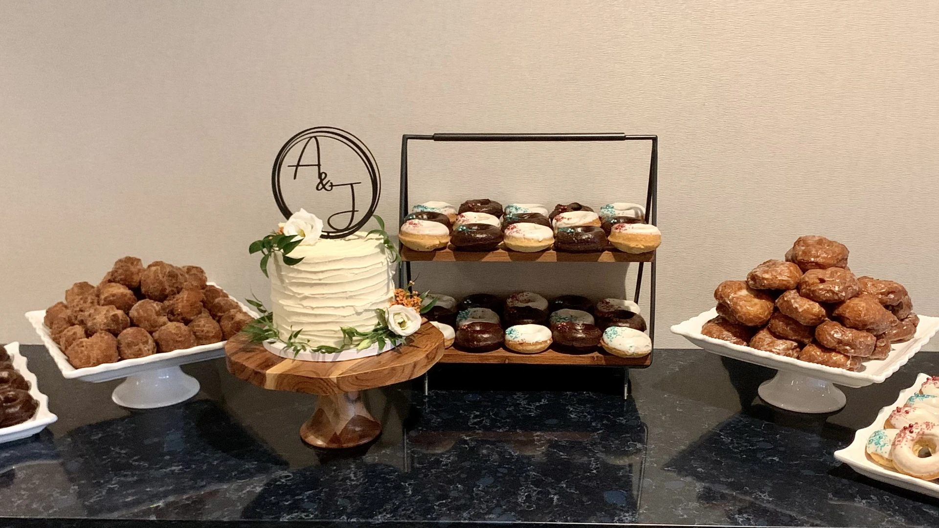A single tier cake with an 'A & J' topper, flanked with trays of donuts.