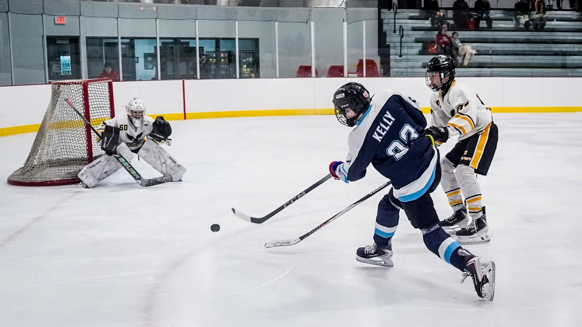 An ice hockey player in a navy blue jersey takes a shot on goal while a defender and goalie prepare to block the puck.