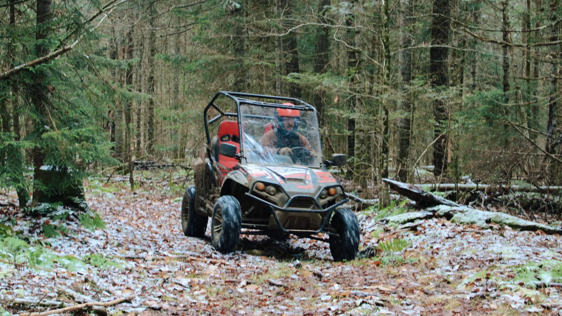 Early snow covers the fallen leaves as a man in a helmet drives a UTV on a path through the trees 