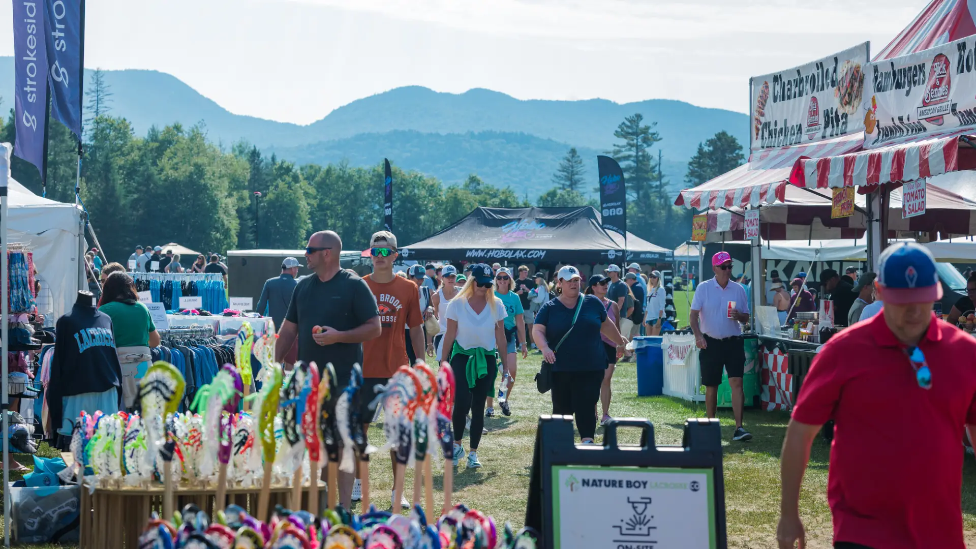 People mill around food and lacrosse gear vendors at Lake Placid Summit Lacrosse Tournament with mountains in background