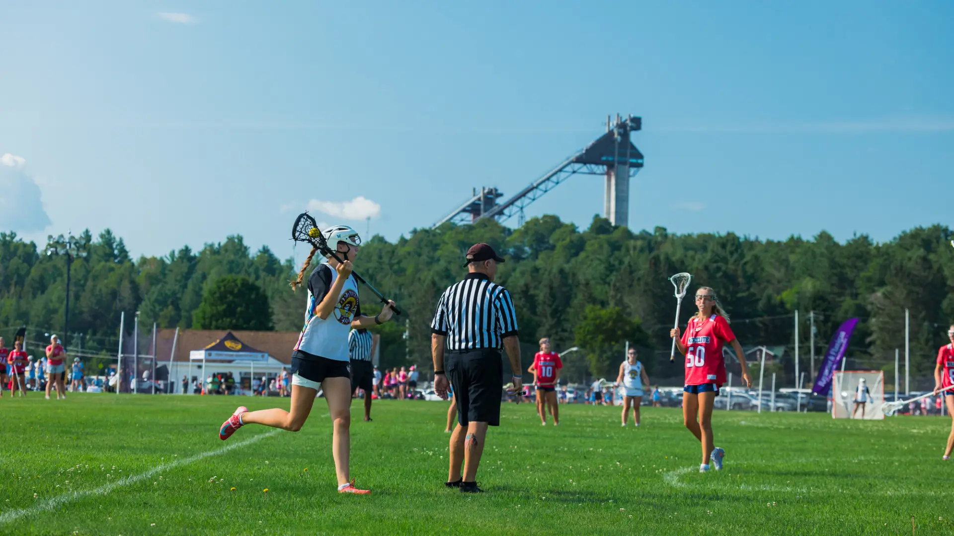 Woman in white and black uniform holds lacrosse ball in stick while running past referee during game on field in Lake Placid