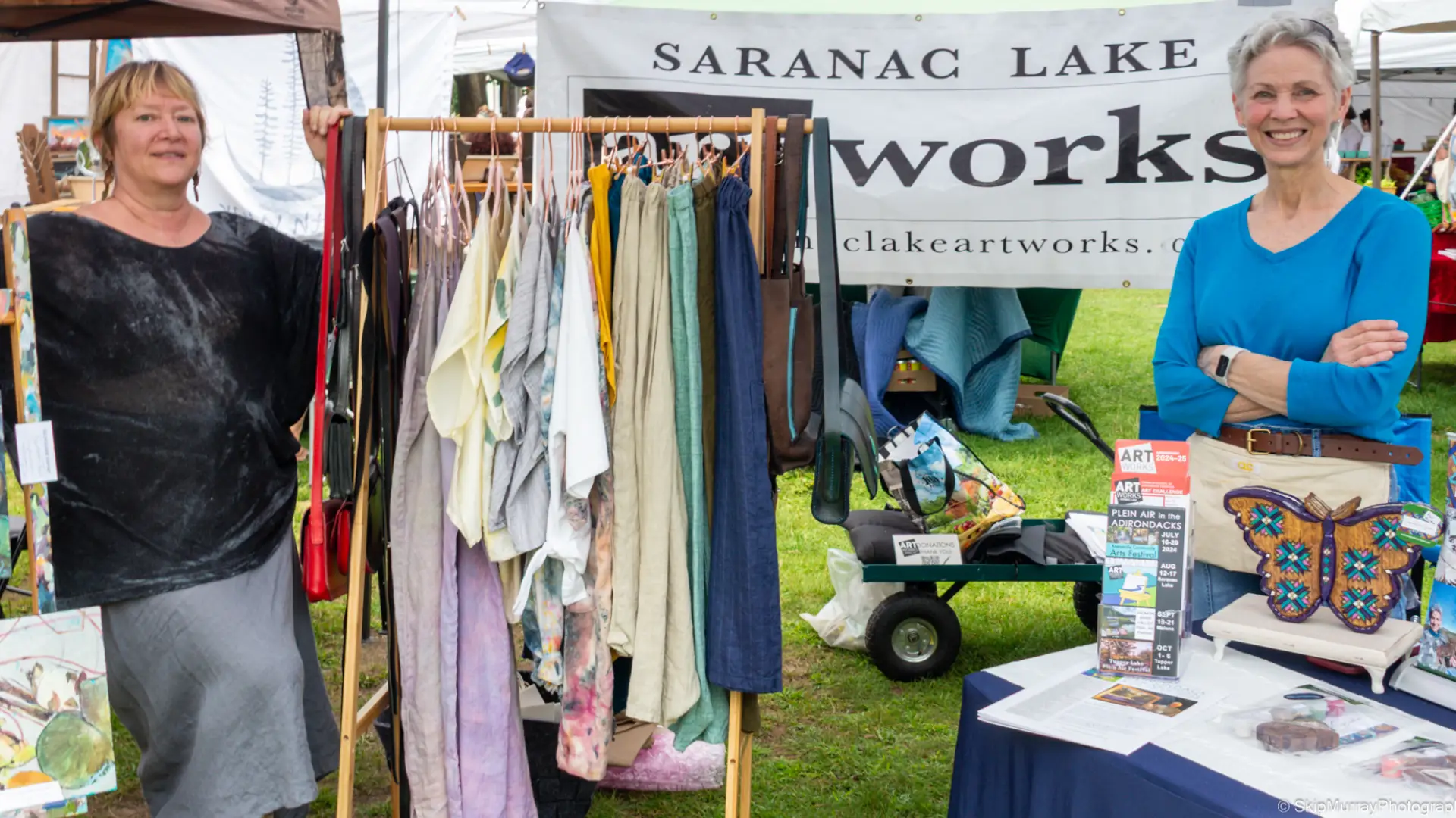 Two women stand by a table and a display rack of clothing outside under a tent with a banner reading Saranac Lake artworks behind them