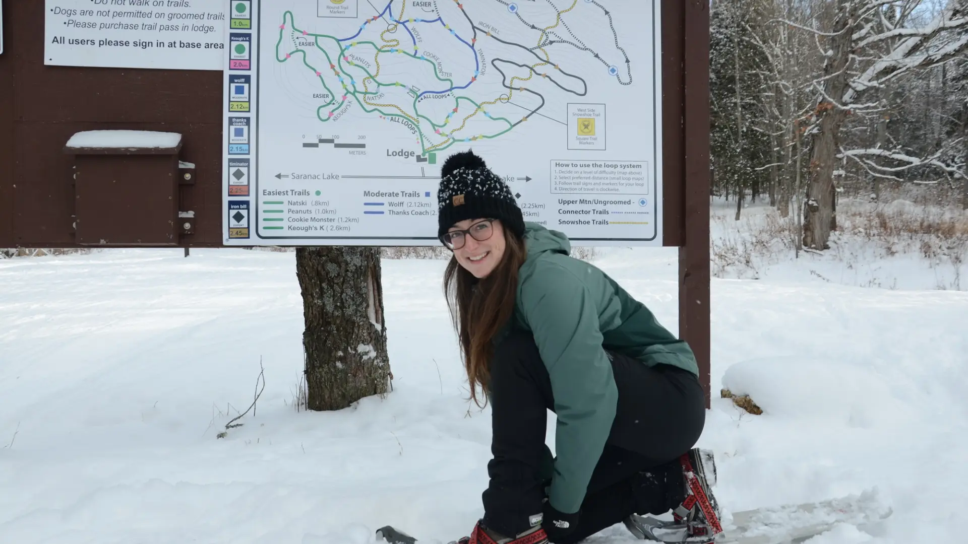Girl smiles at the camera as she tightens her snowshoes in front of a Dewey Mountain sign