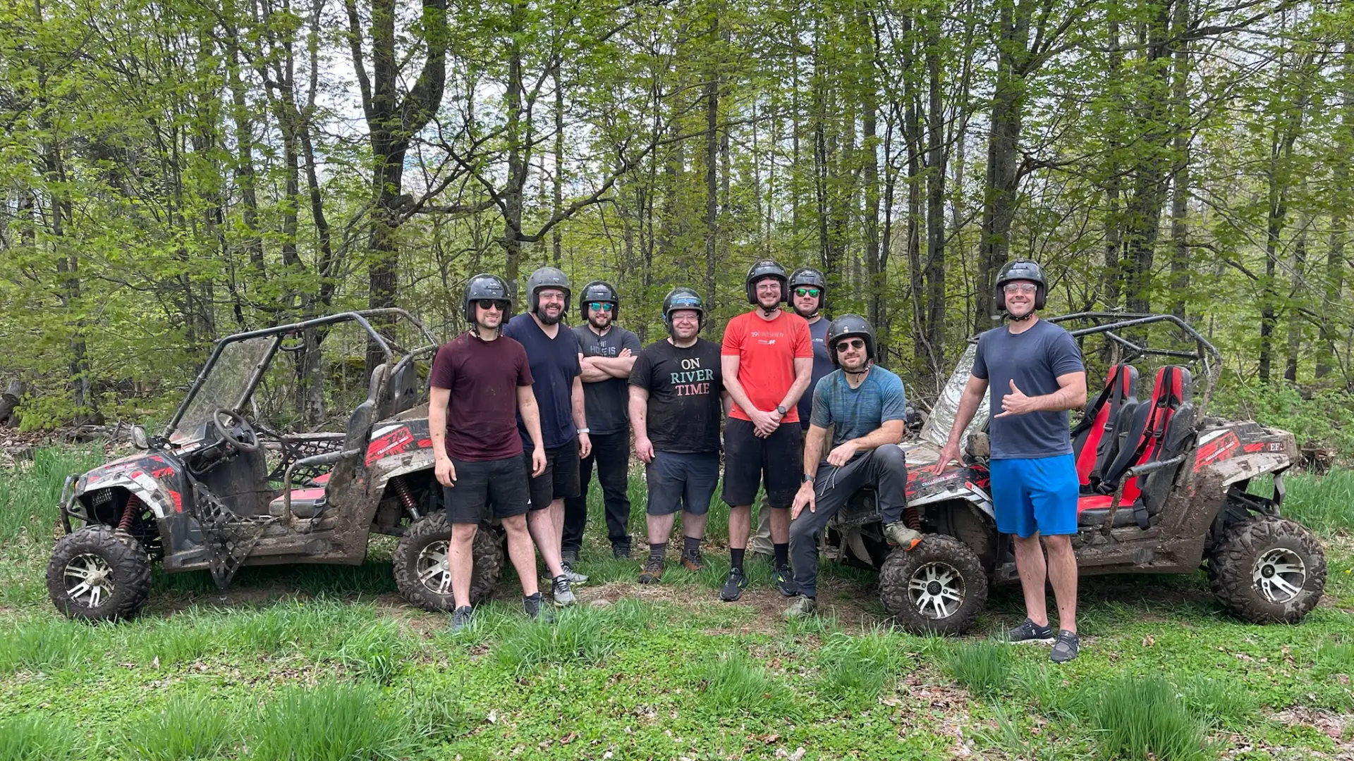Eight happy young men stand together wearing helmets and smiling between all terrain vehicles in a grassy patch