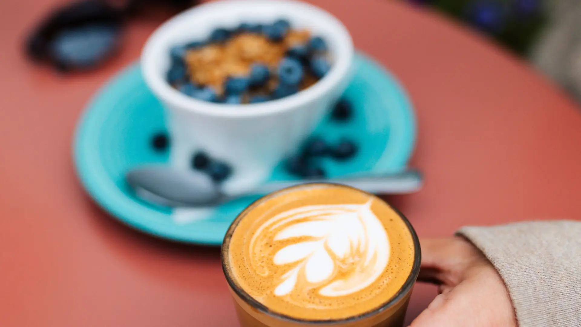 A hand holds a latte on a table next to a breakfast of berries and granola over yogurt.
