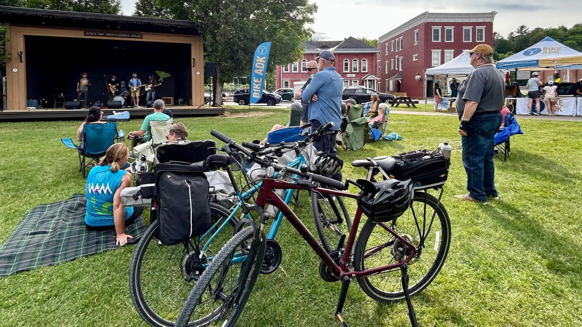 A group of adults with bicycles and picnic blankets gather on the green lawn of a village park.