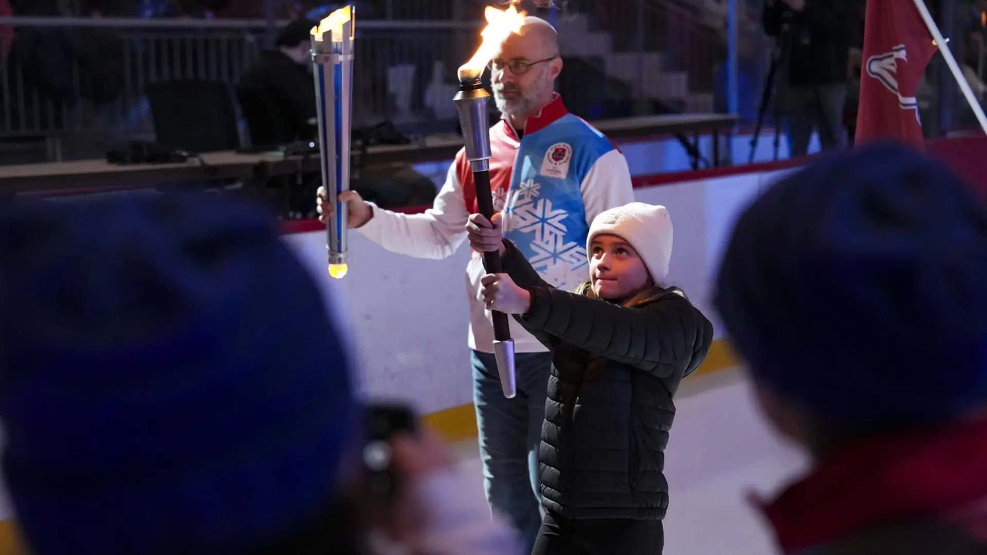 A young girl in a white beanie holds a flaming torch, assisted by an older man in a blue and red jacket. 