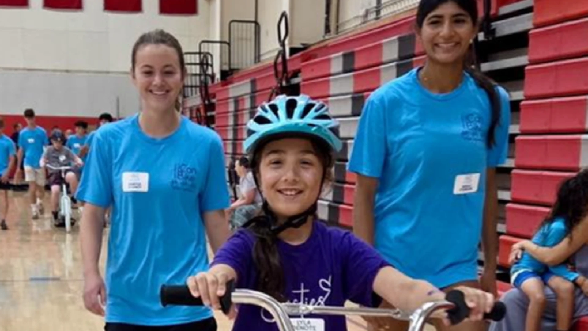 A child with blue helmet smiles on a bike in a gymnasium flanked by two volunteers.