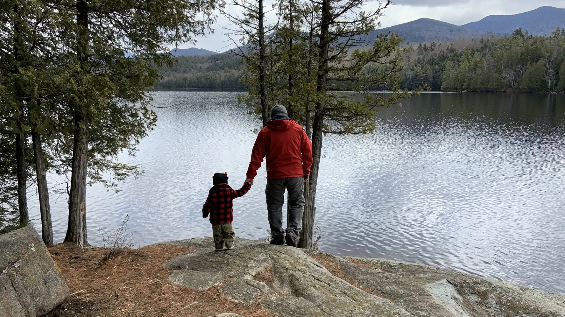 Father and son hold hands as they walk over a rock near the shoreline of Moose Pond