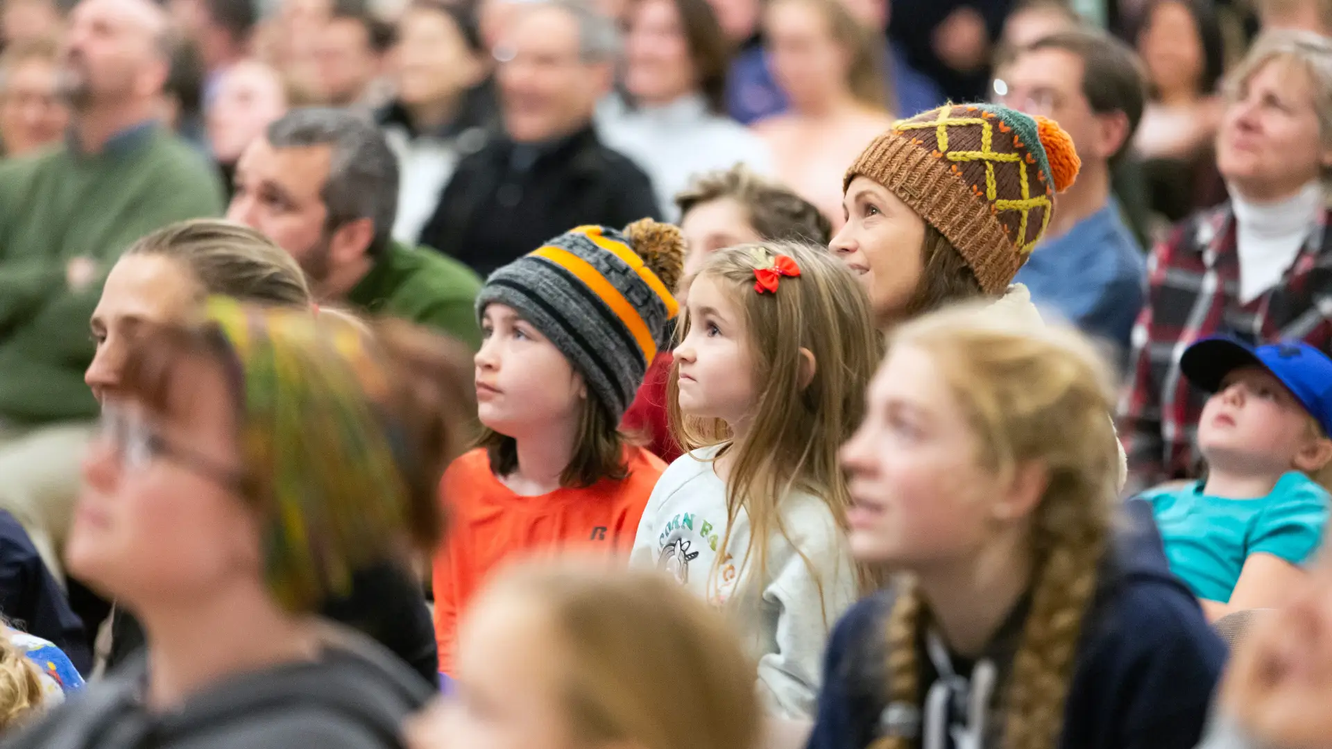 A group of people, including children and adults, attentively listen in an indoor setting.