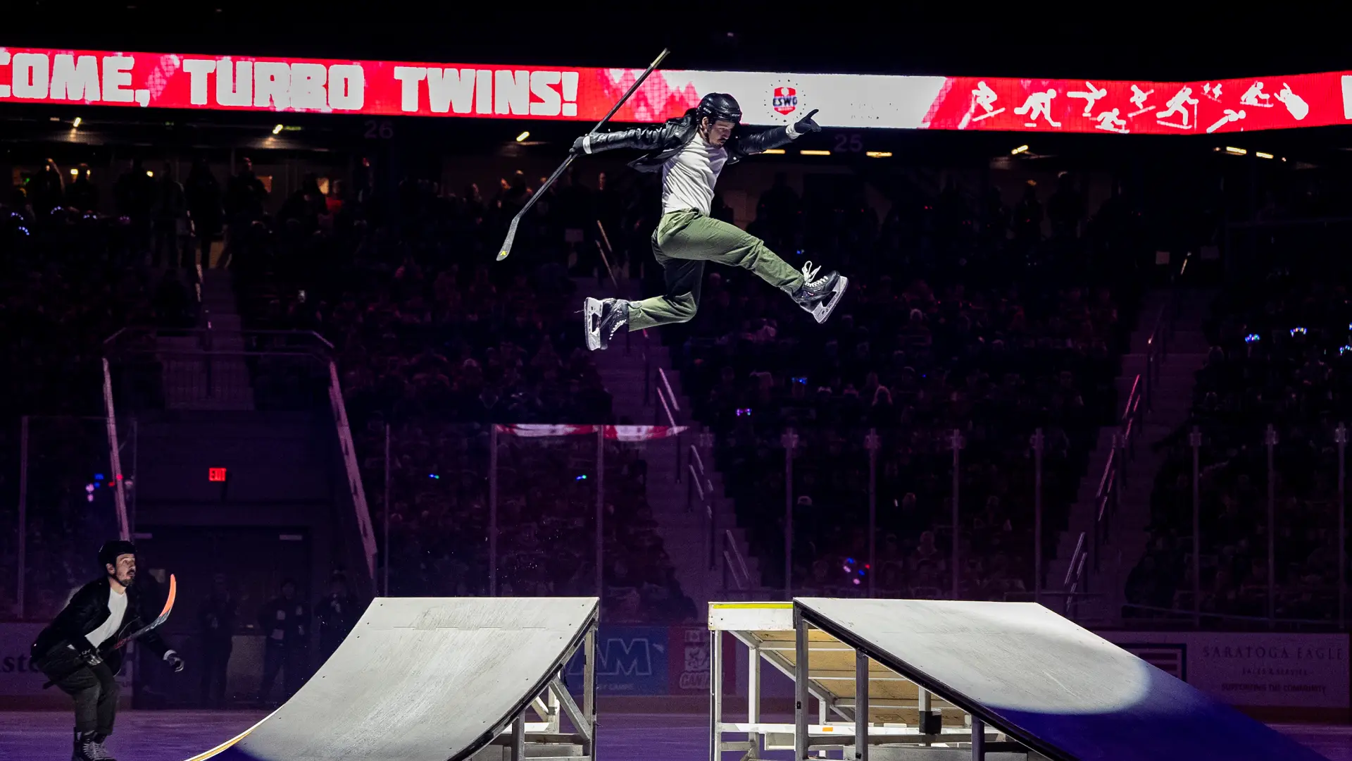 An ice skater performs a high jump over a ramp on an ice rink, holding a hockey stick as part of a stunt performance.