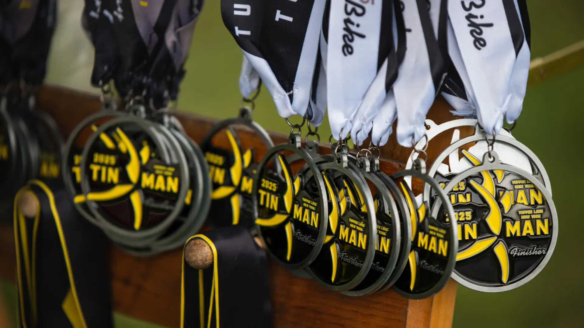 A row of silver 2025 Tupper Lake Tinman finisher medals with yellow and black accents hang from ribbons on a wooden rack.