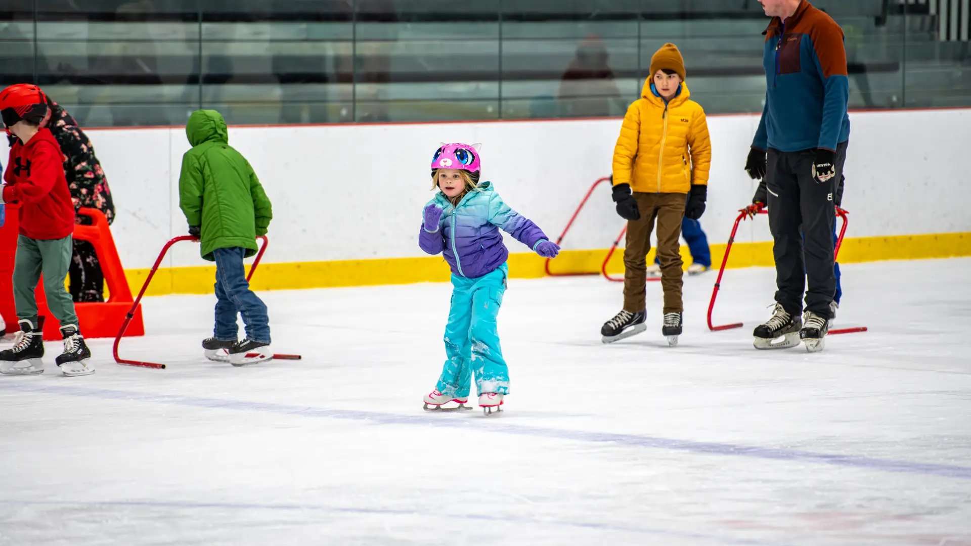 A child in a bright outfit skates without training bar while other still use them in the background.