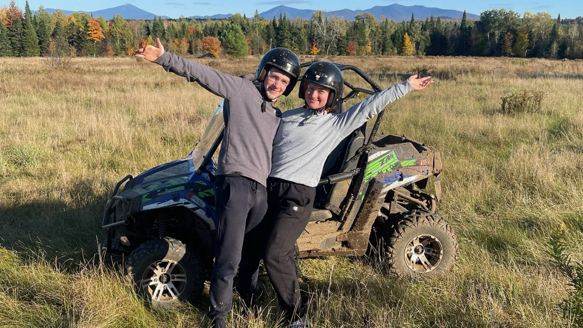 A happy couple smile into the camera with an arm raised in excitement in front of a UTV