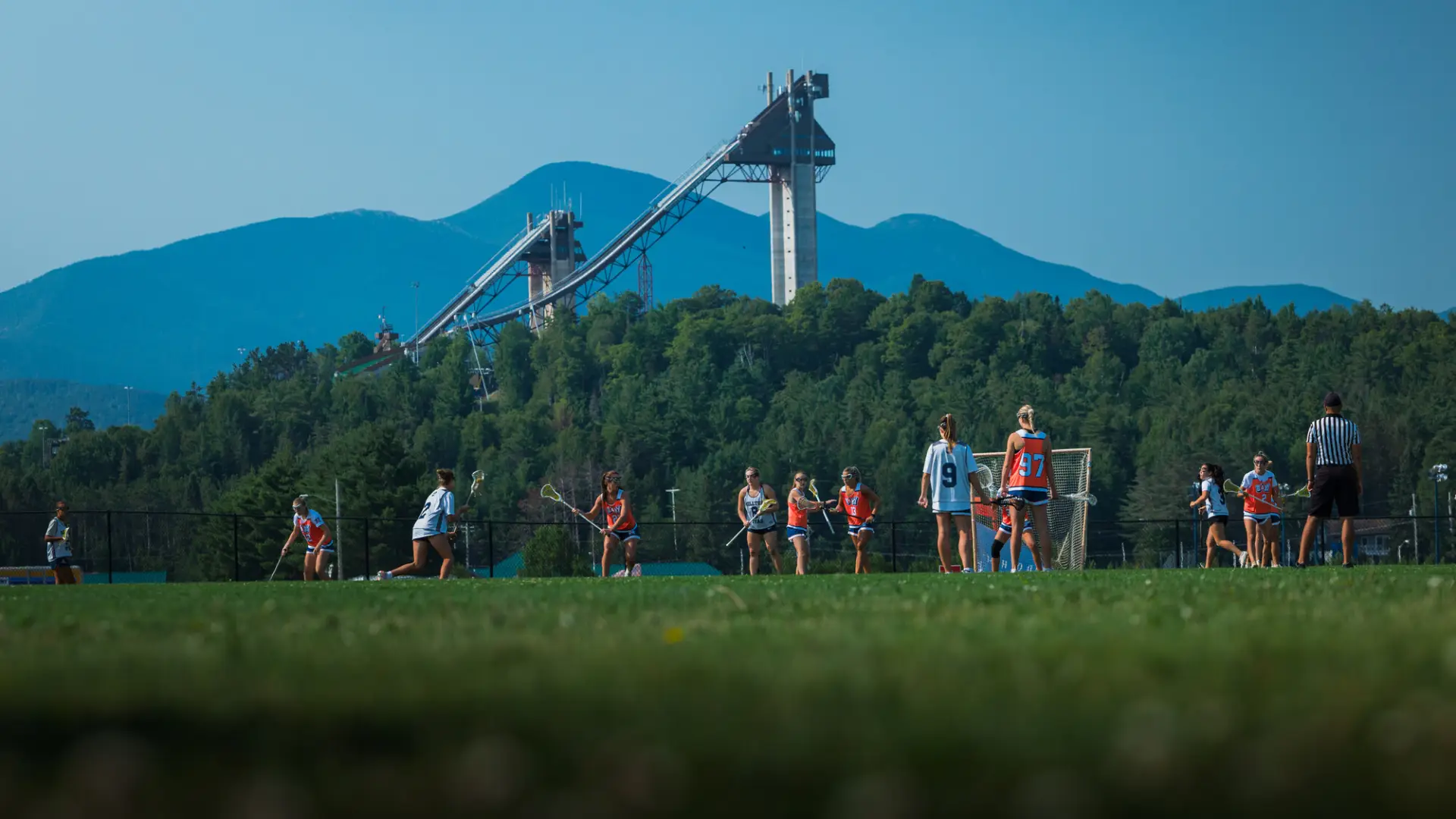 Female lacrosse players in white jerseys play against team in orange jerseys on field in front of mountains and ski jumps