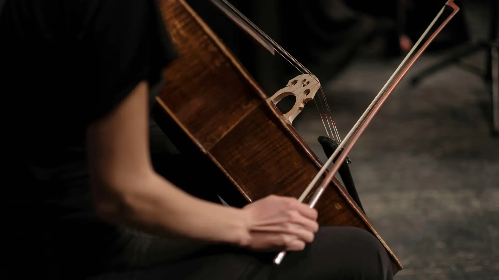 Person in black sits while holding cello in one hand an a bow in the other