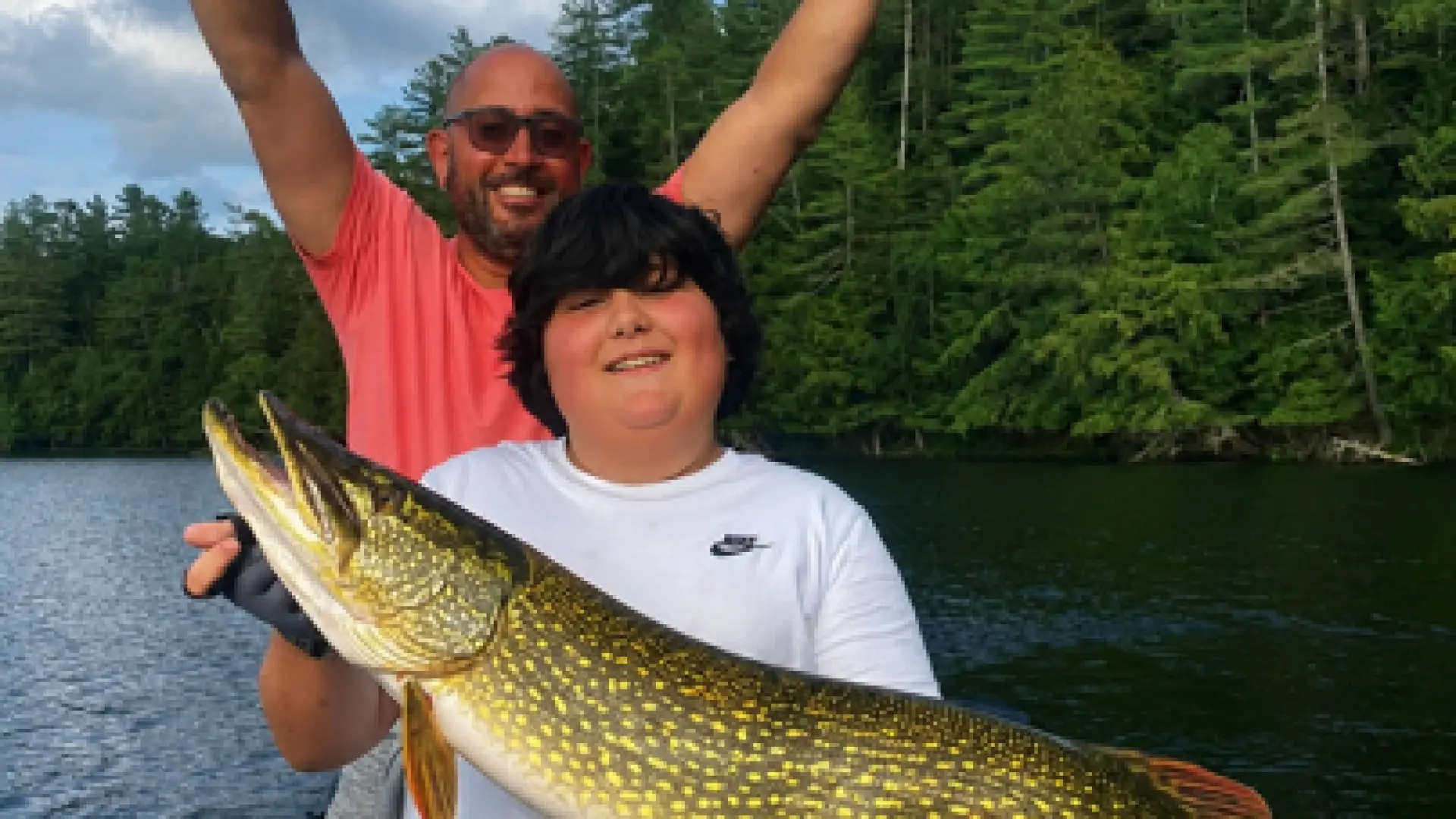 A kid holds a large pike while a man throws up his hands in victory behind.