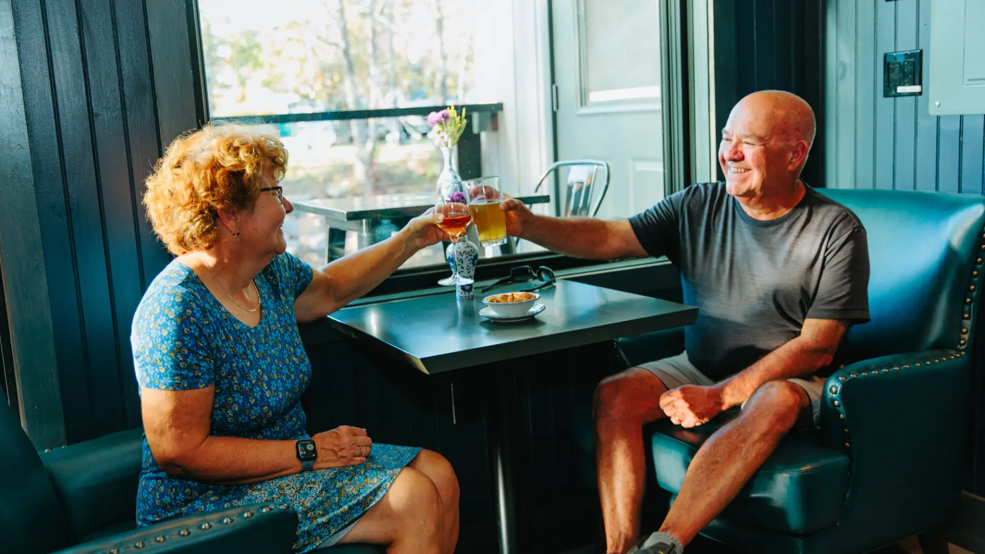 A smiling man and woman sit in plush leather armchairs by a window, clinking a wine glass and a beer glass over a table.